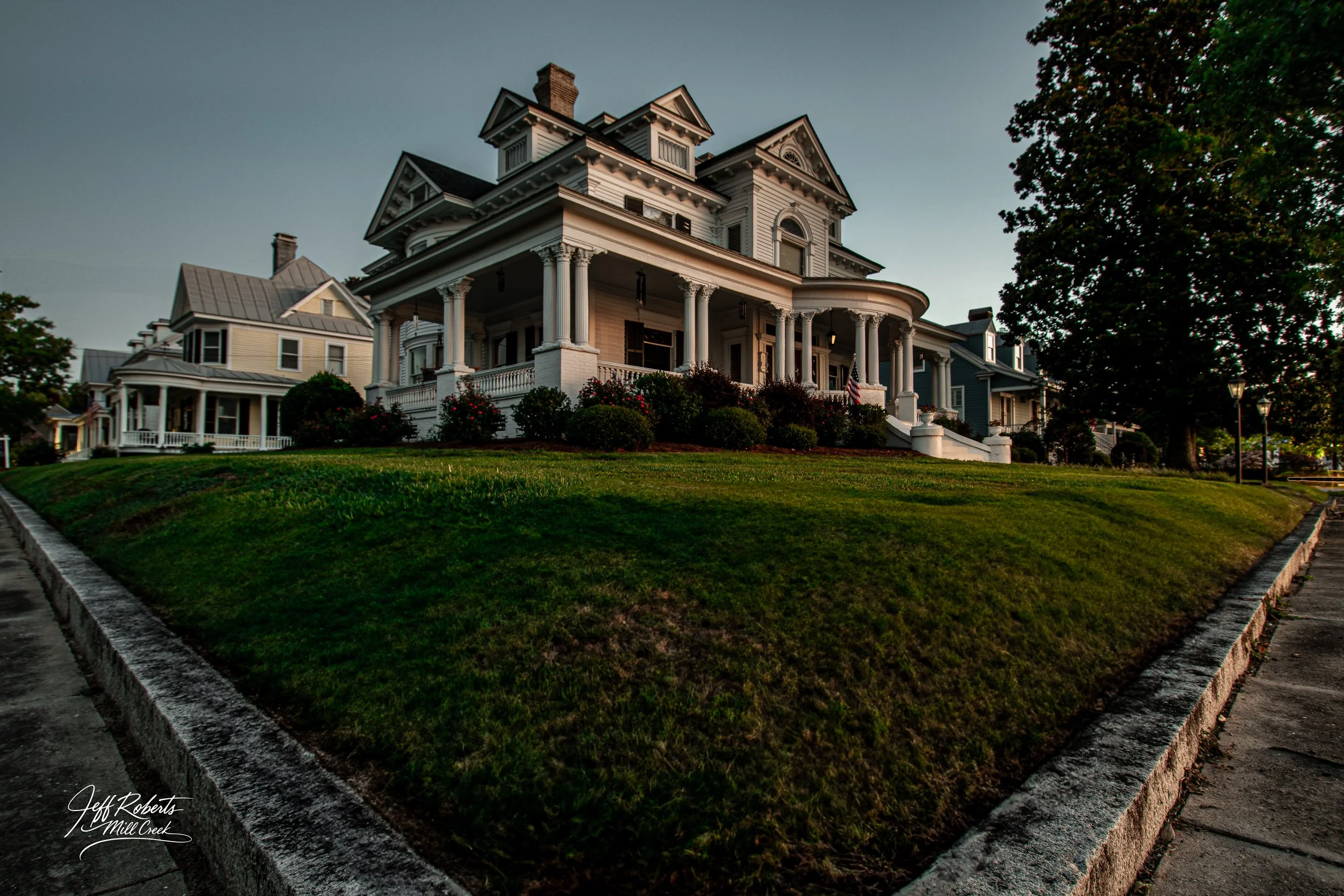 A large, historic Victorian-style house on a hill, featuring a wide front porch with columns, surrounded by a grassy lawn, trees, and a sidewalk.