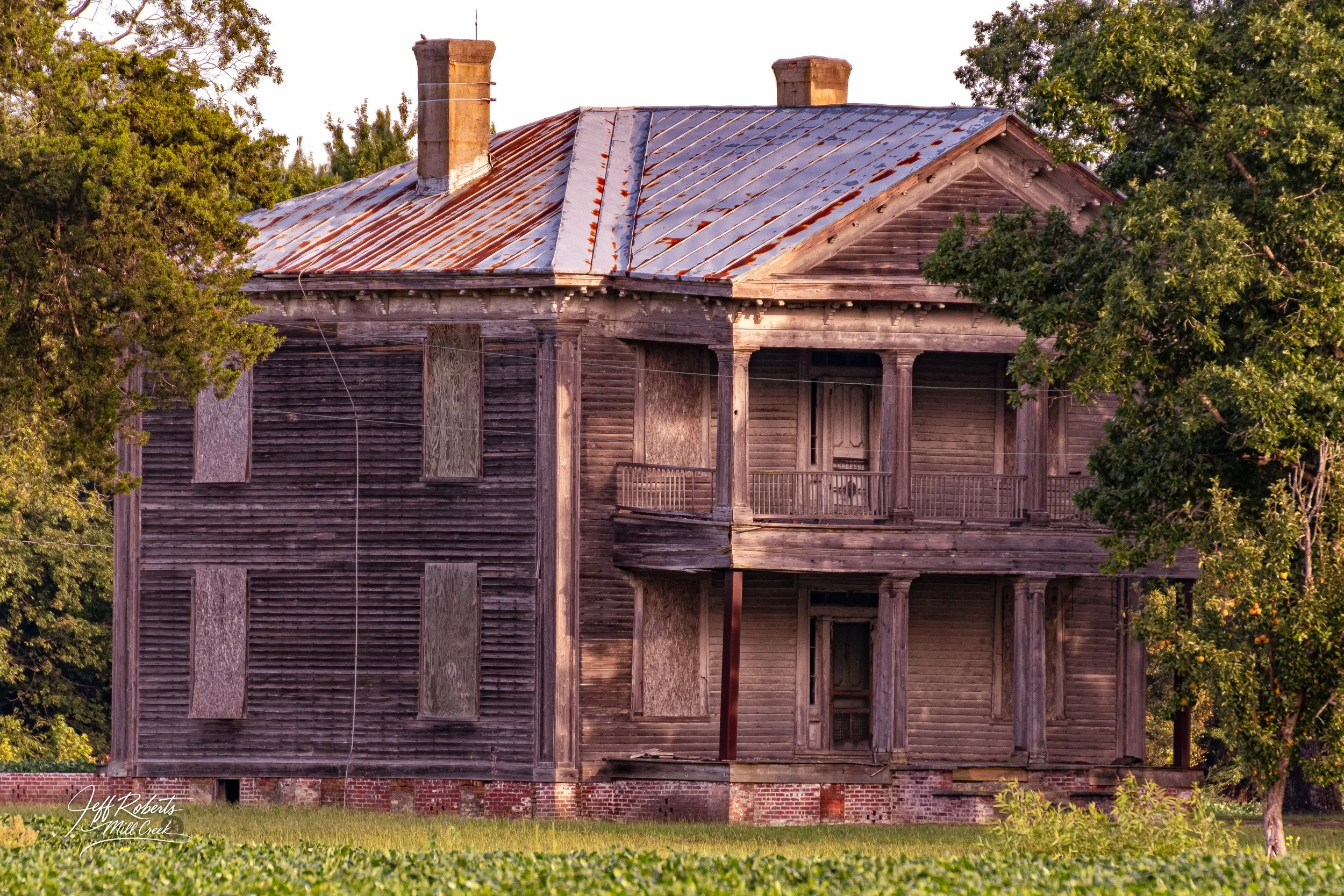 An old, abandoned two-story wooden house with boarded-up windows and a rusted metal roof, surrounded by trees and grass.