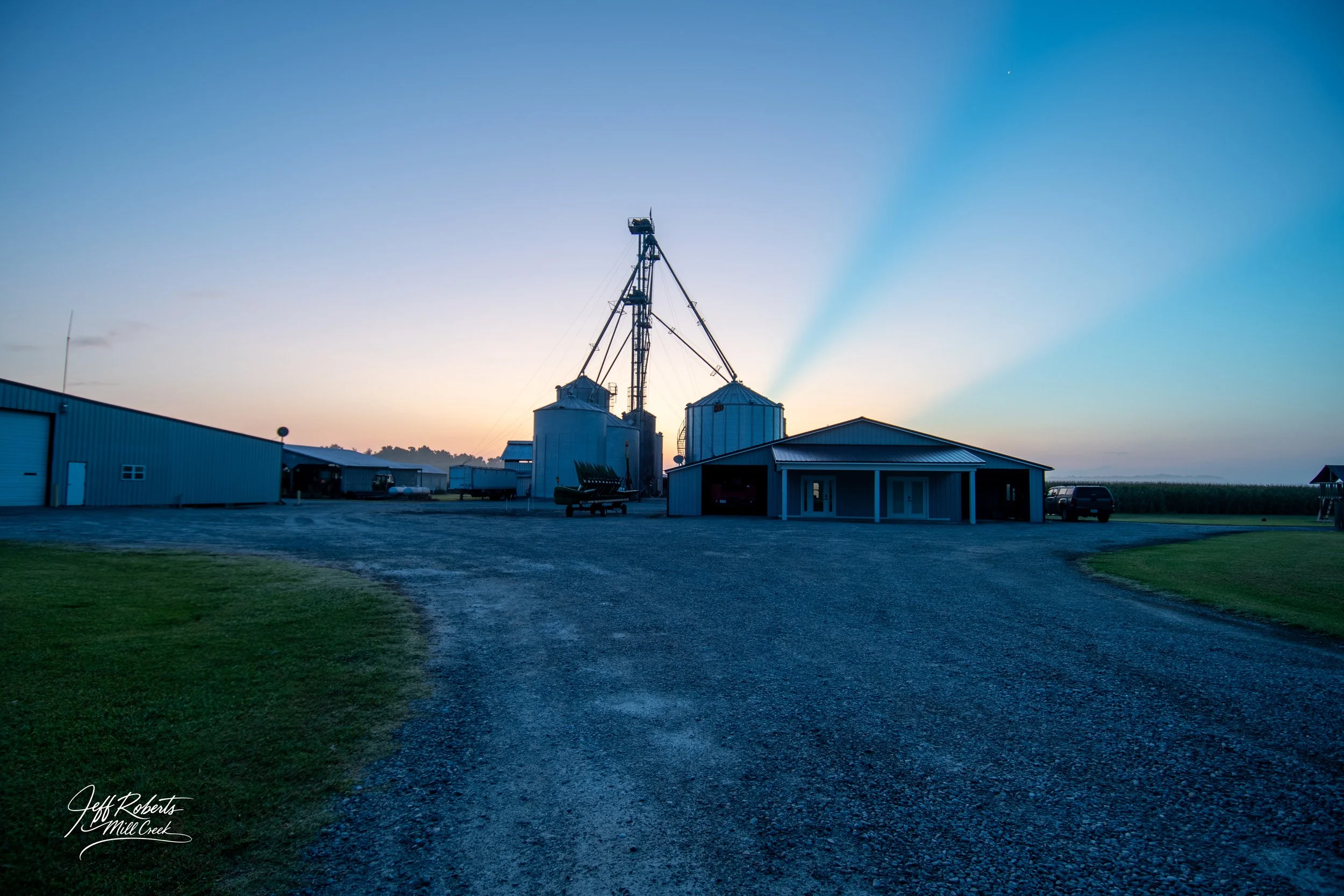 Farm buildings including silos and storage sheds at sunset, with gravel driveway and grassy areas.