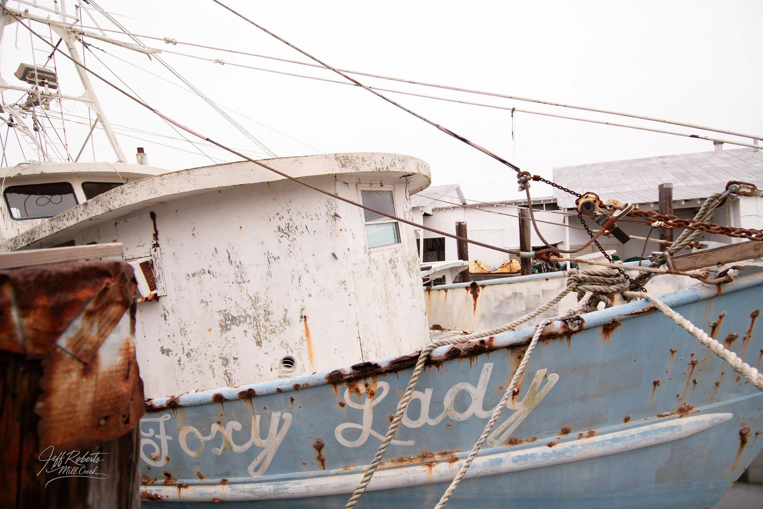 An old, rusted boat named 'Foxy Lady' docked at a pier, with rust spots and peeling paint, surrounded by weathered ropes and wooden posts.