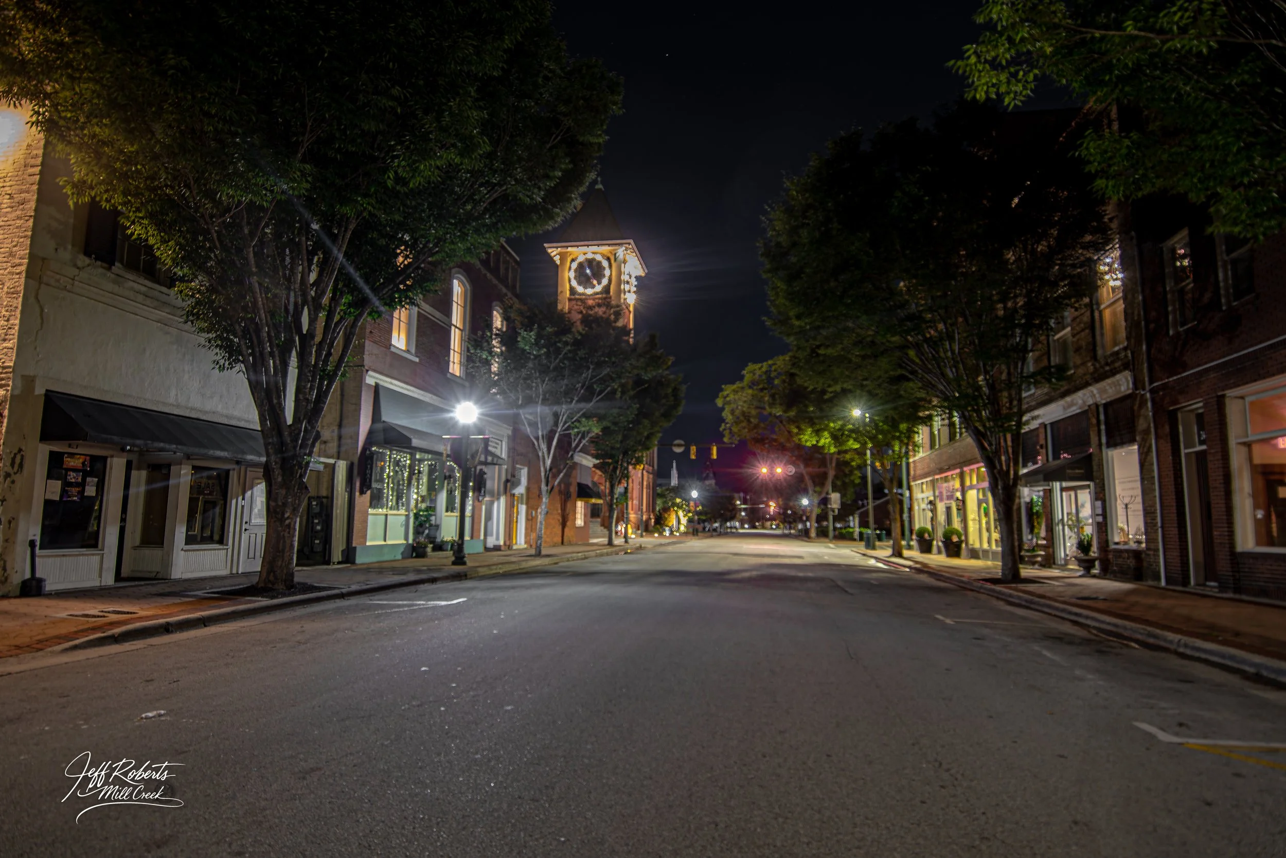 Empty downtown street at night with trees lining the sides, buildings with storefronts, and a clock tower with a clock face illuminated in the background.