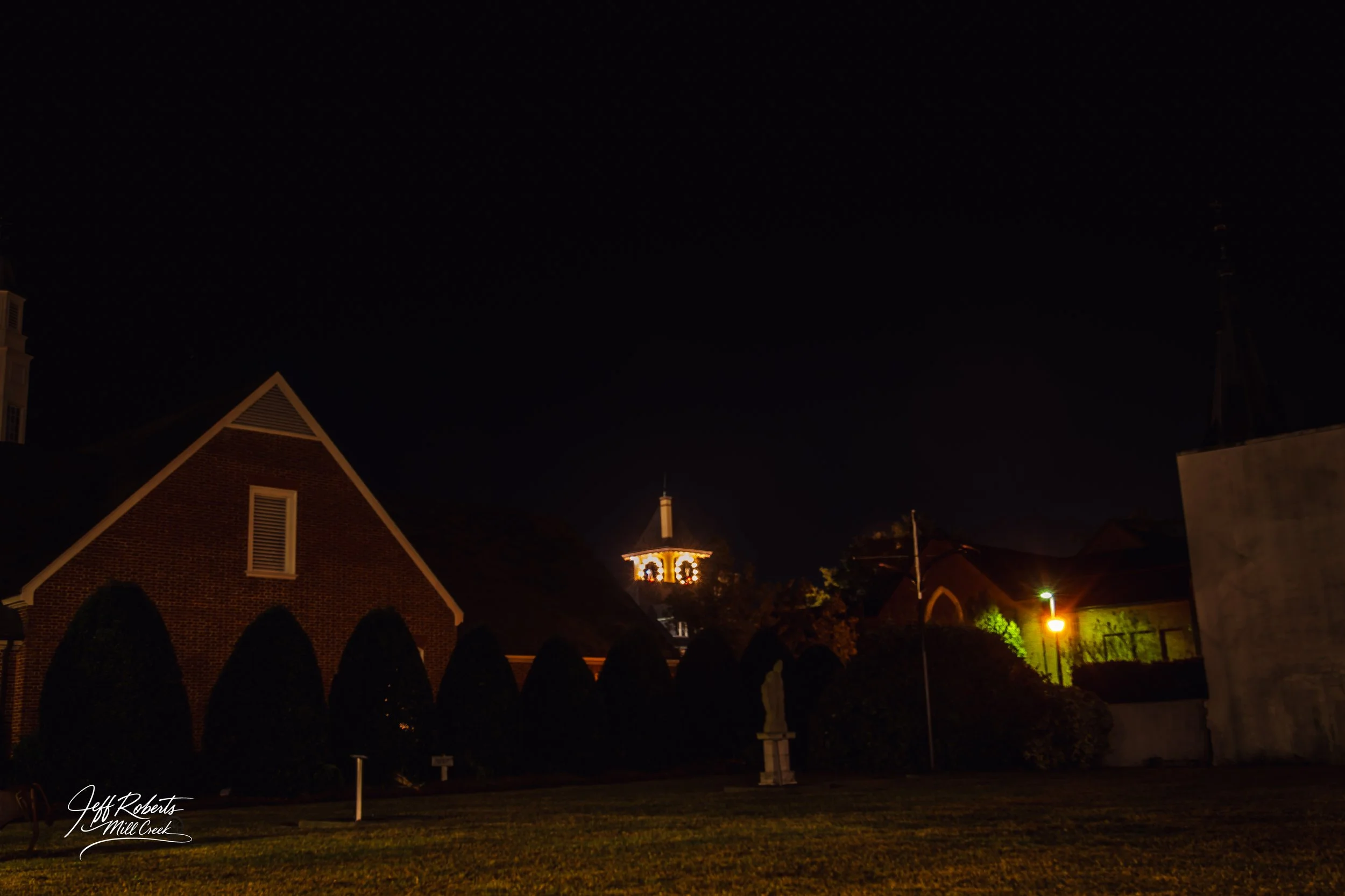 Nighttime street scene with illuminated church steeple, brick building, trees, and sidewalk.