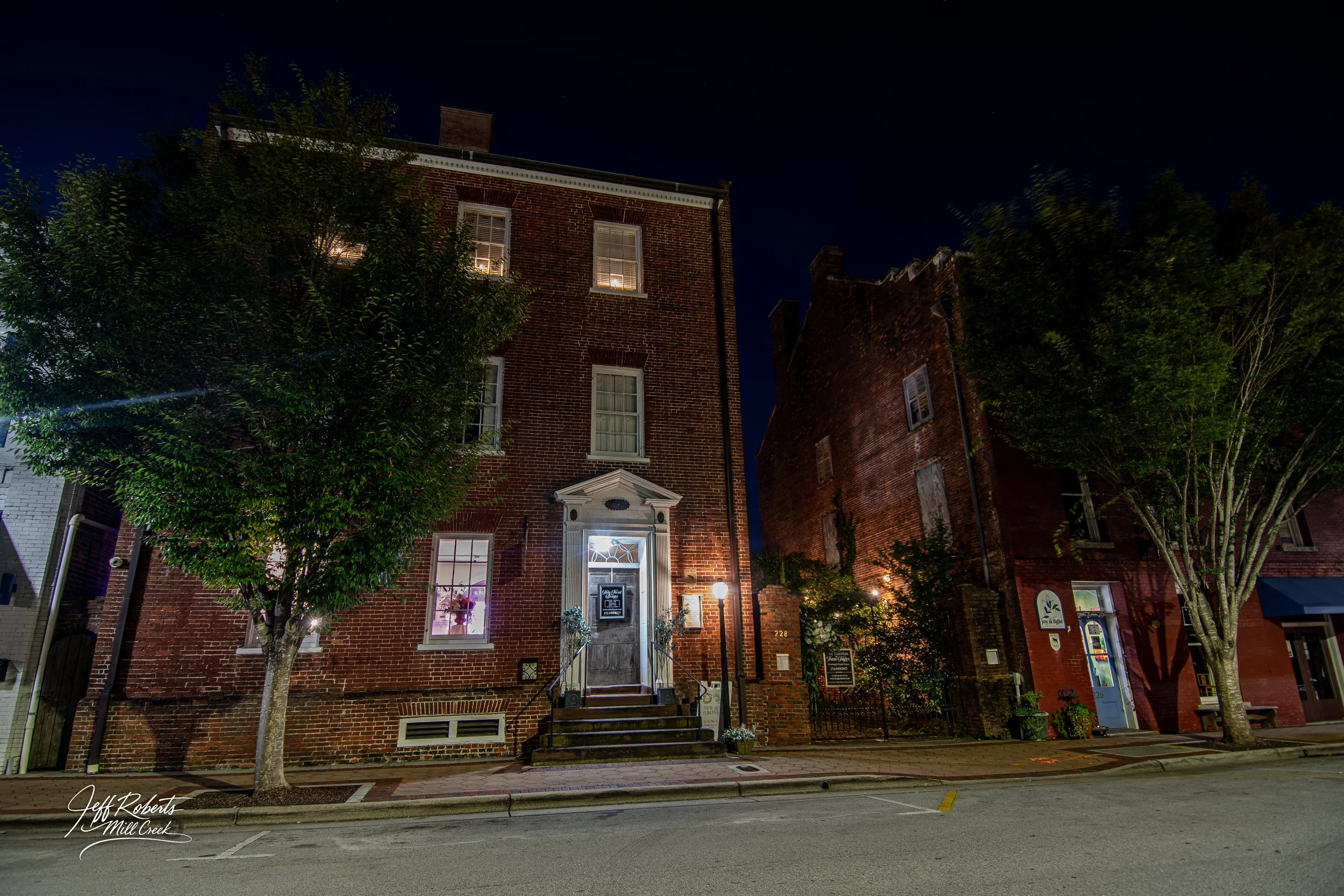 Night view of brick buildings with lit windows, trees, and a sidewalk in a downtown area.