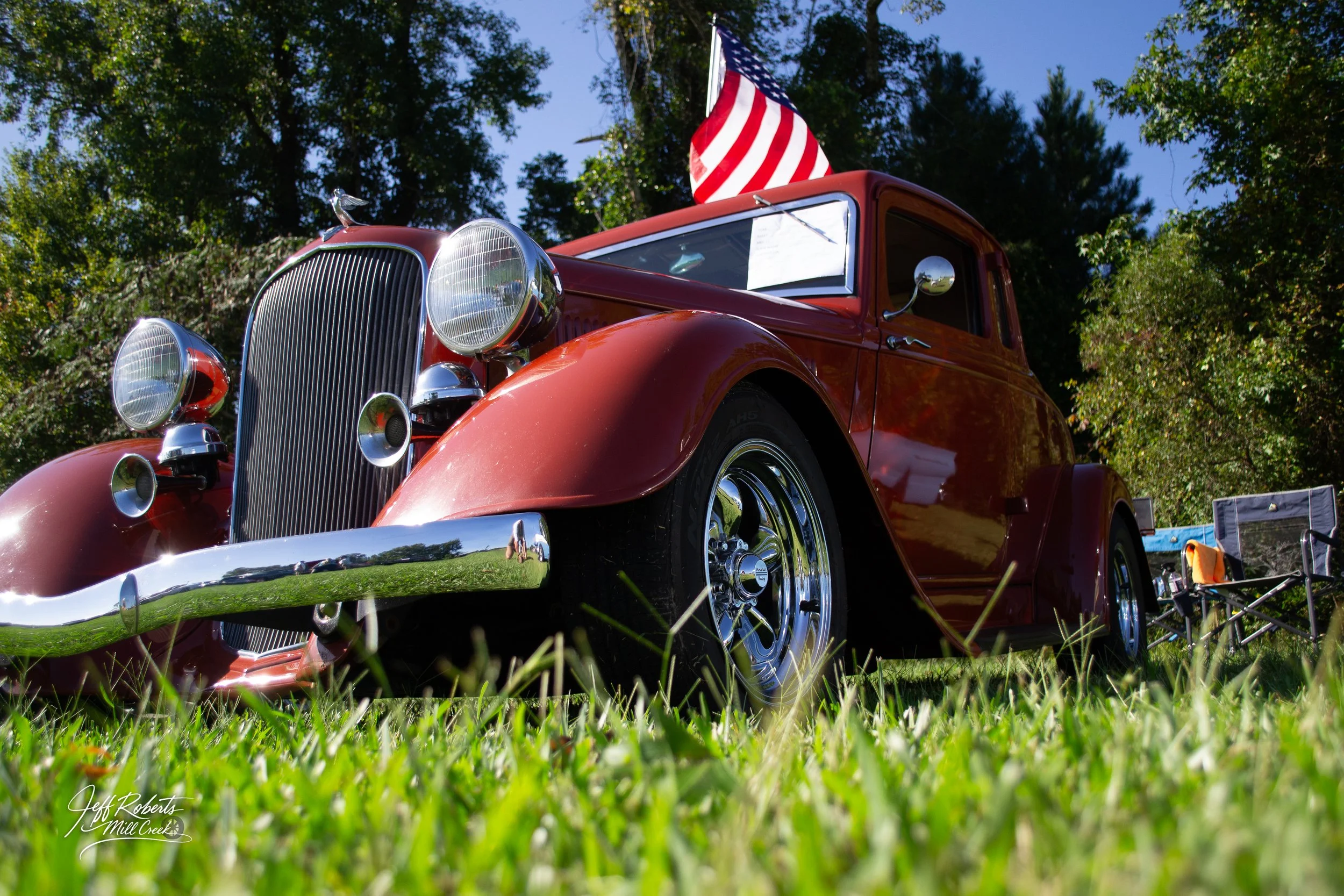 A vintage red car with a shiny chrome bumper and large round headlights parked on grass, with an American flag attached to its window, surrounded by trees during daytime.