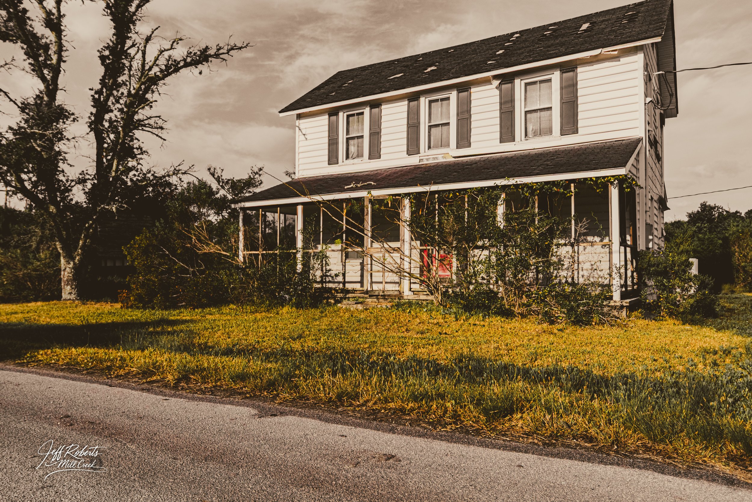 An old white two-story house with black shutters, a front porch overgrown with bushes, and a large tree on the left side, under a cloudy sky.