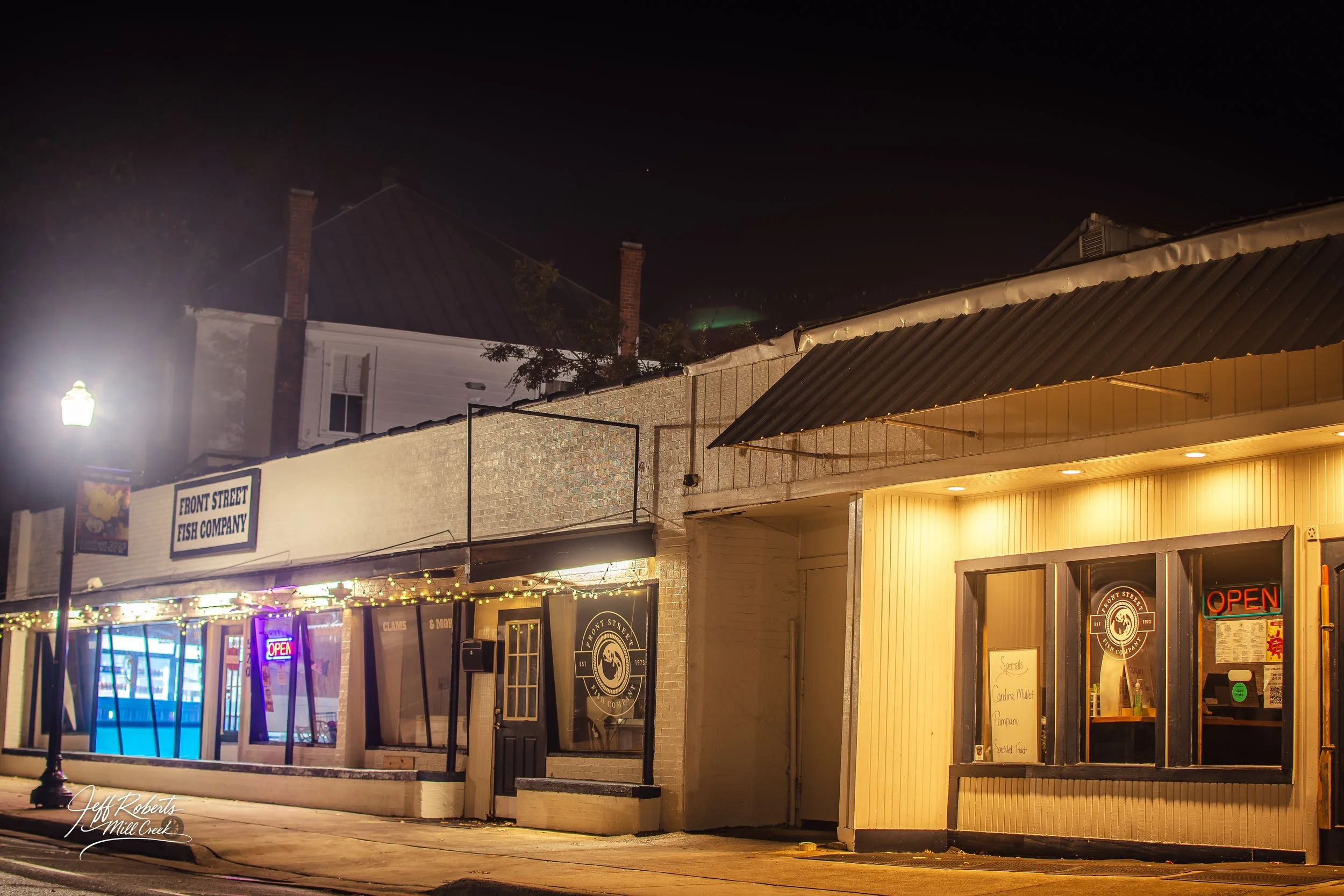 Night view of a small storefront called Front Street Fish Company with illuminated signs, one says 'Open'. The building has a light-colored exterior with large windows and a small sidewalk in front. A street lamp is visible on the left side.