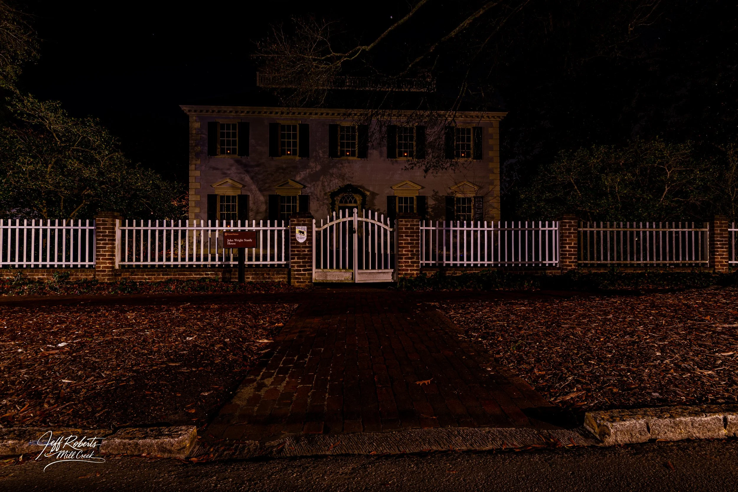 Night view of historic house behind a white picket fence, with brick pathway leading to the gate, surrounded by trees and fallen leaves.