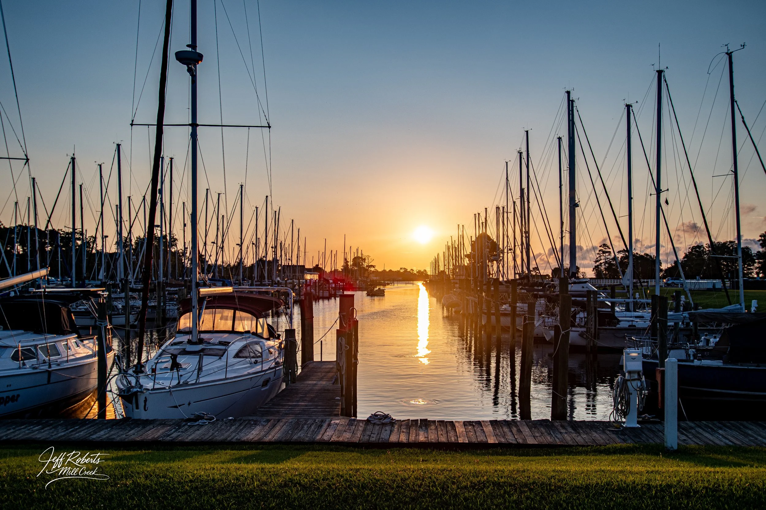 Sunset over a marina with sailboats docked on both sides and their reflections in the water.