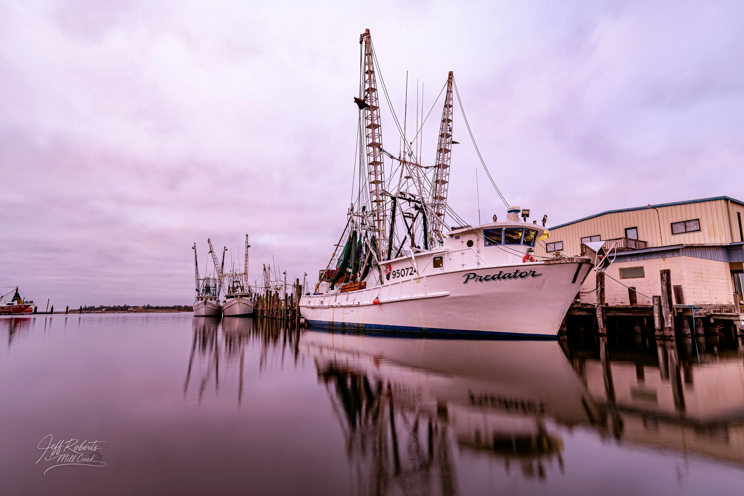 Fishing boats docked at a marina on calm water with cloudy sky and a building on the dock