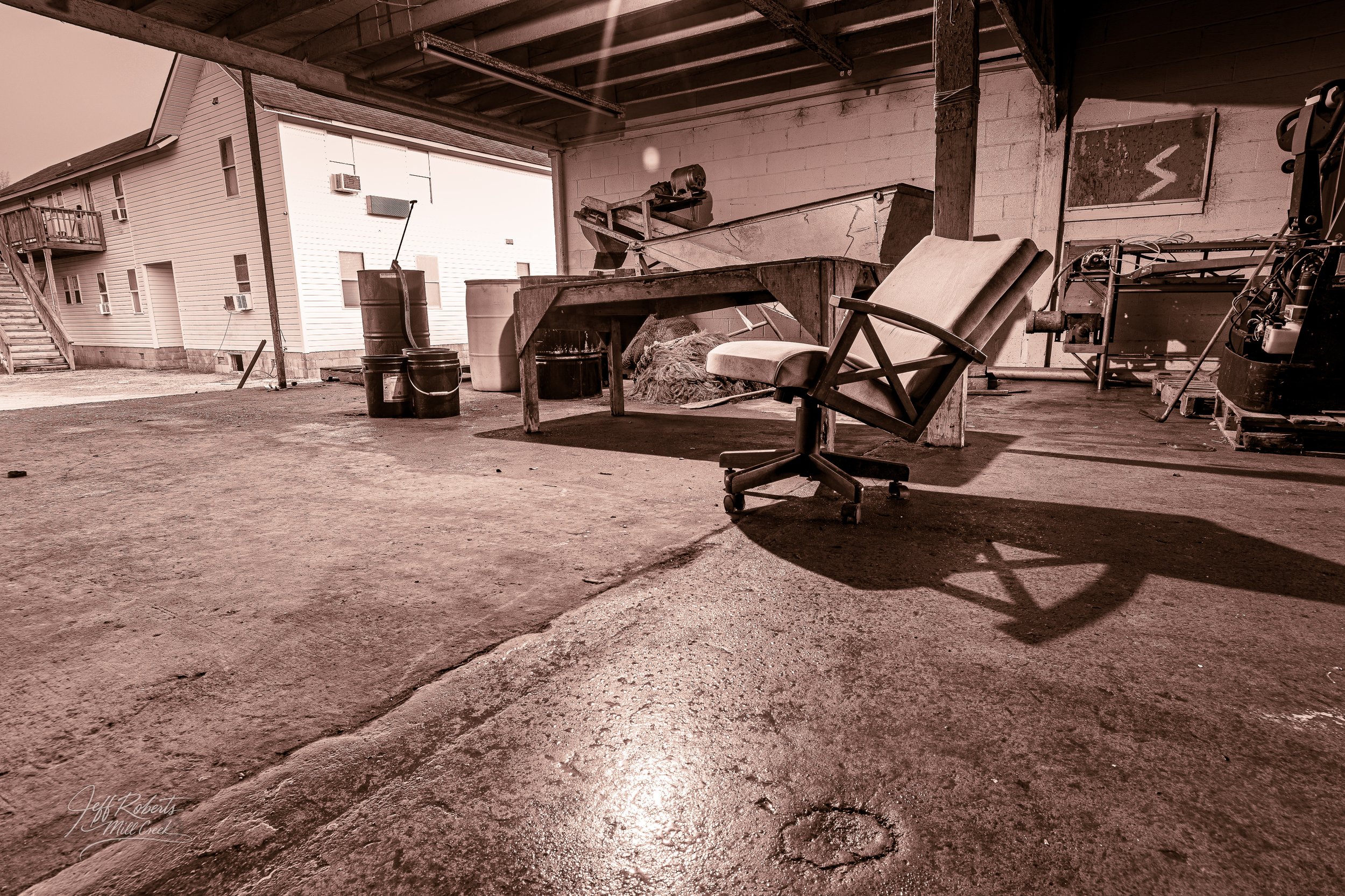 An empty industrial or workshop space with a worn office chair, a wooden table, and various barrels and equipment. The area has concrete floors and is partially open to the outside.