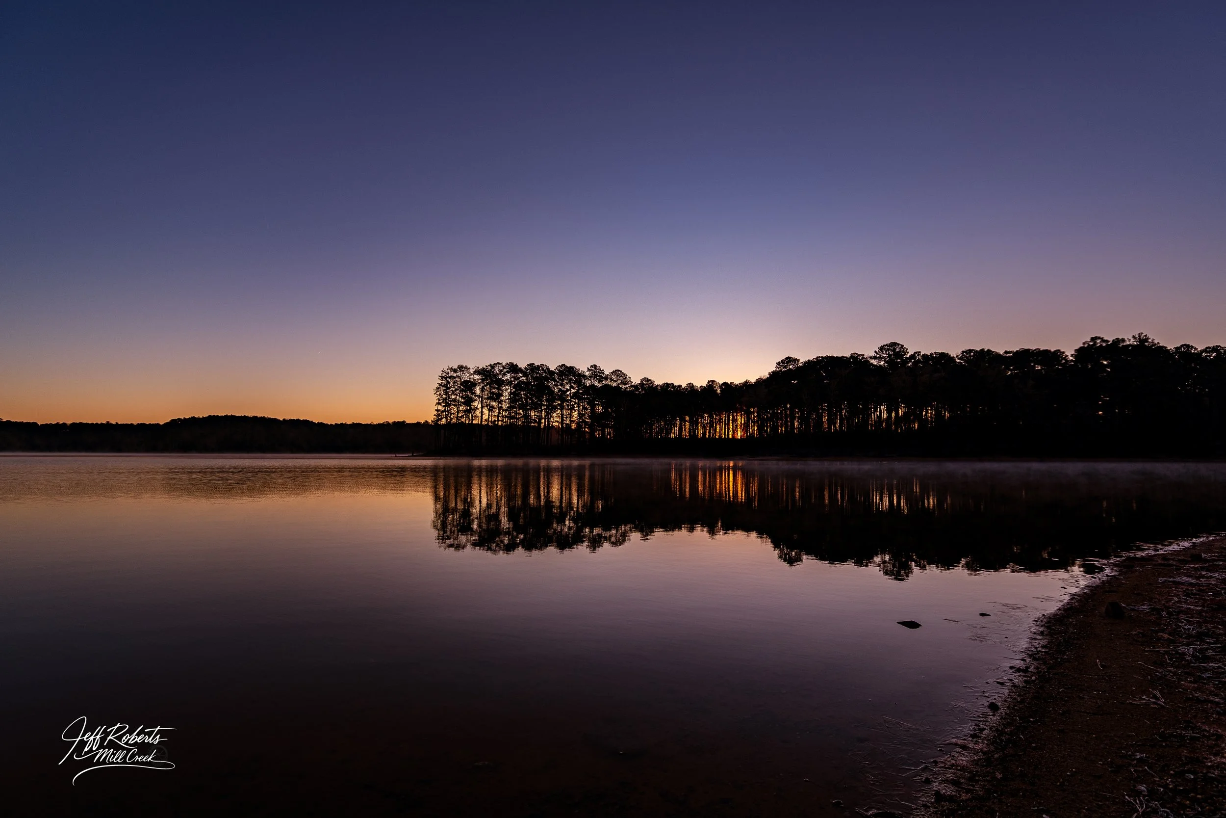 Dusk over a calm lake with a silhouetted tree line reflection, a slight fog on the water, and a fading sunset sky.