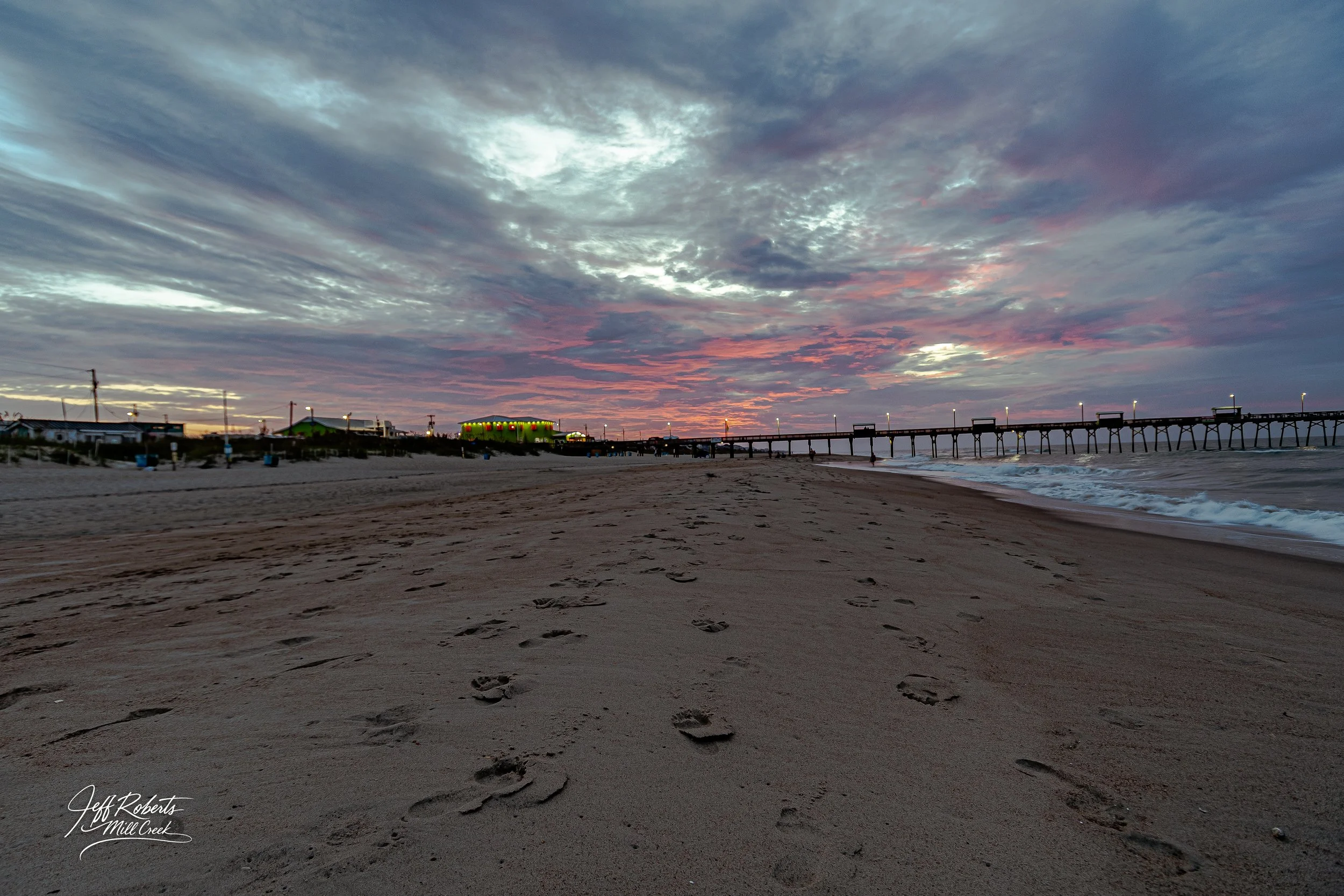 Footprints in the sand on a beach at sunset with a pier extending into the water and colorful clouds in the sky.