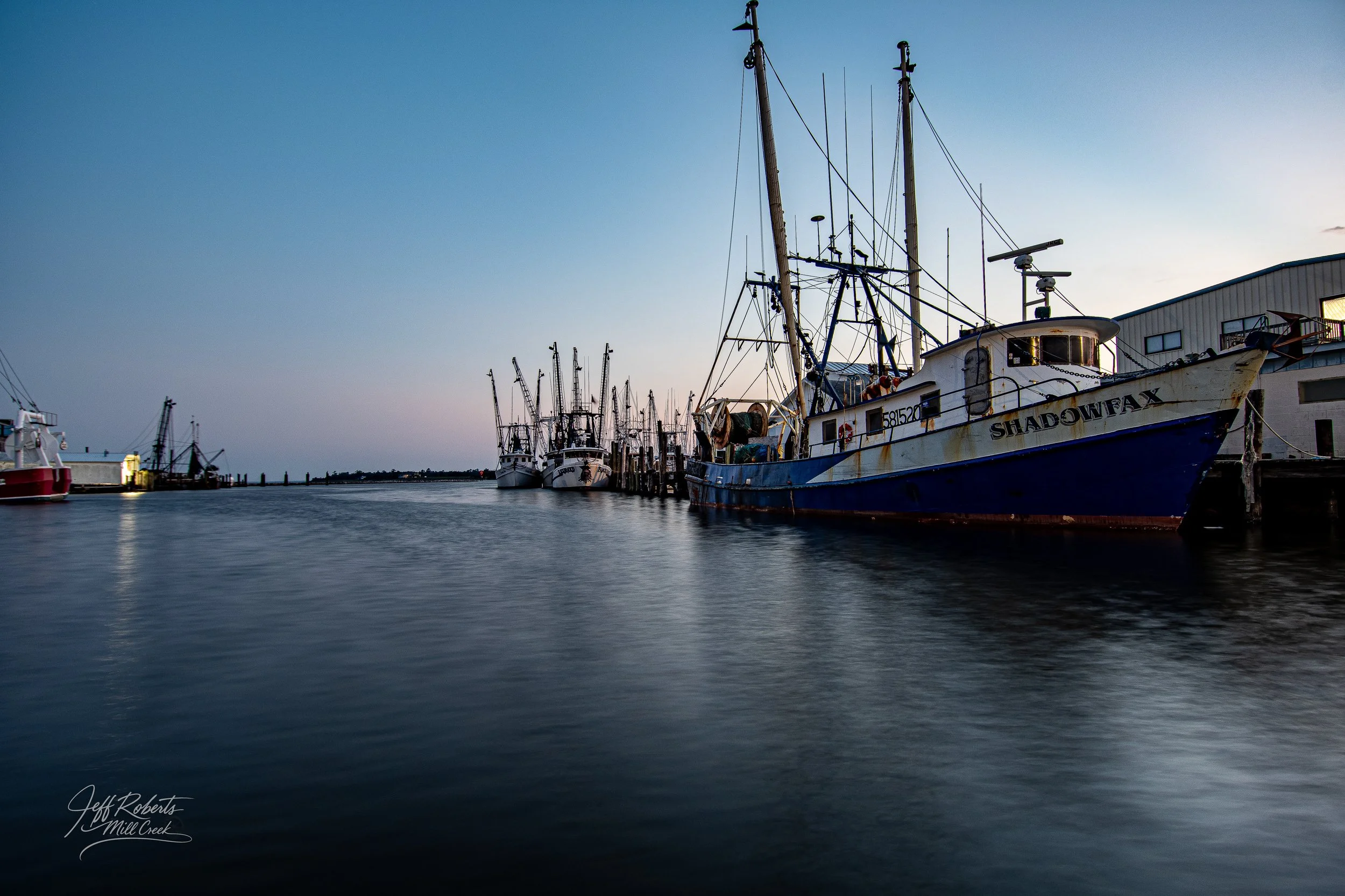 Docked fishing boats at a harbor during dusk, with calm water reflecting the sky and boats.