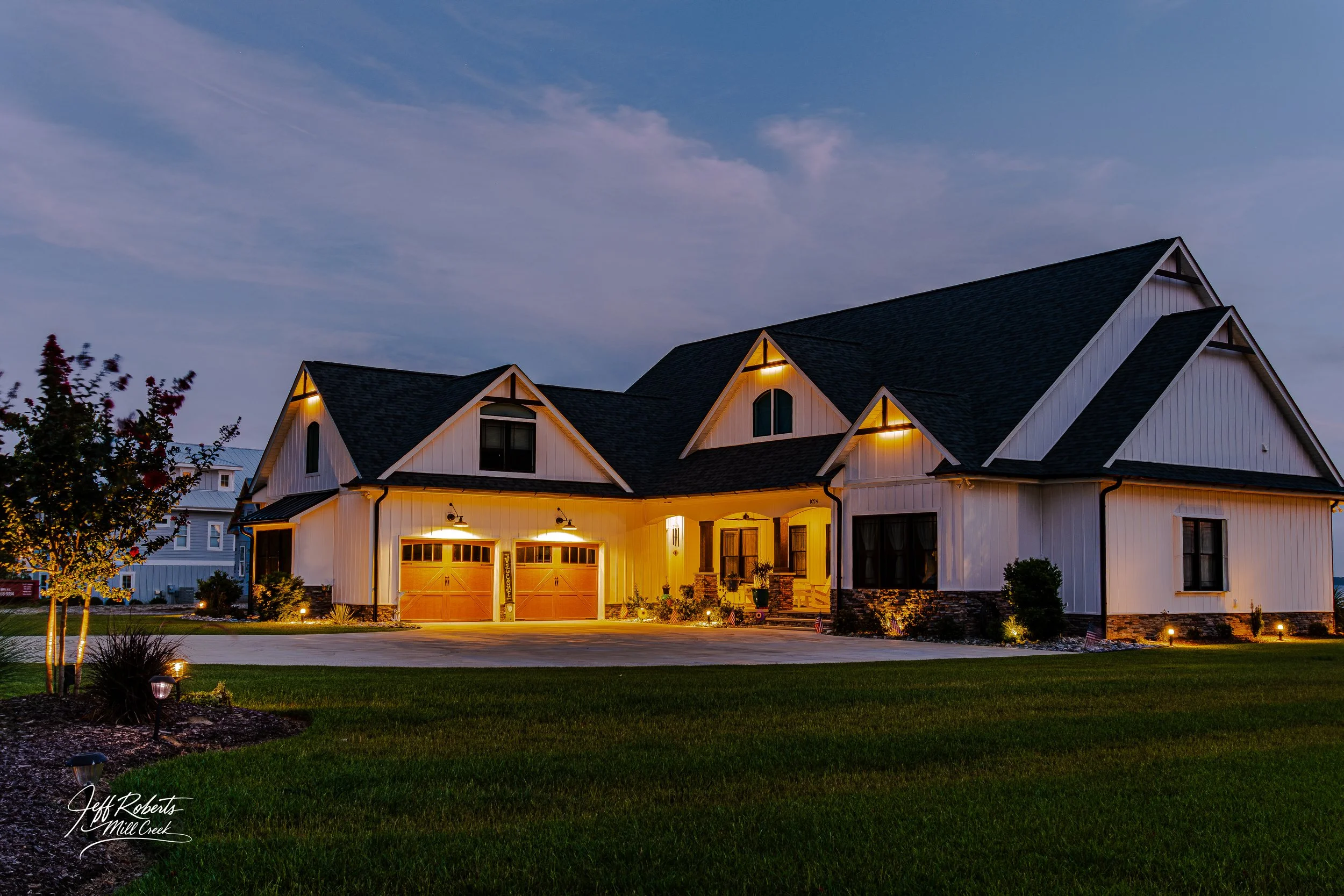 A large two-story house with white exterior siding, black roof, and wooden garage doors, illuminated at dusk with outdoor lights, surrounded by a well-maintained lawn and landscaped garden.