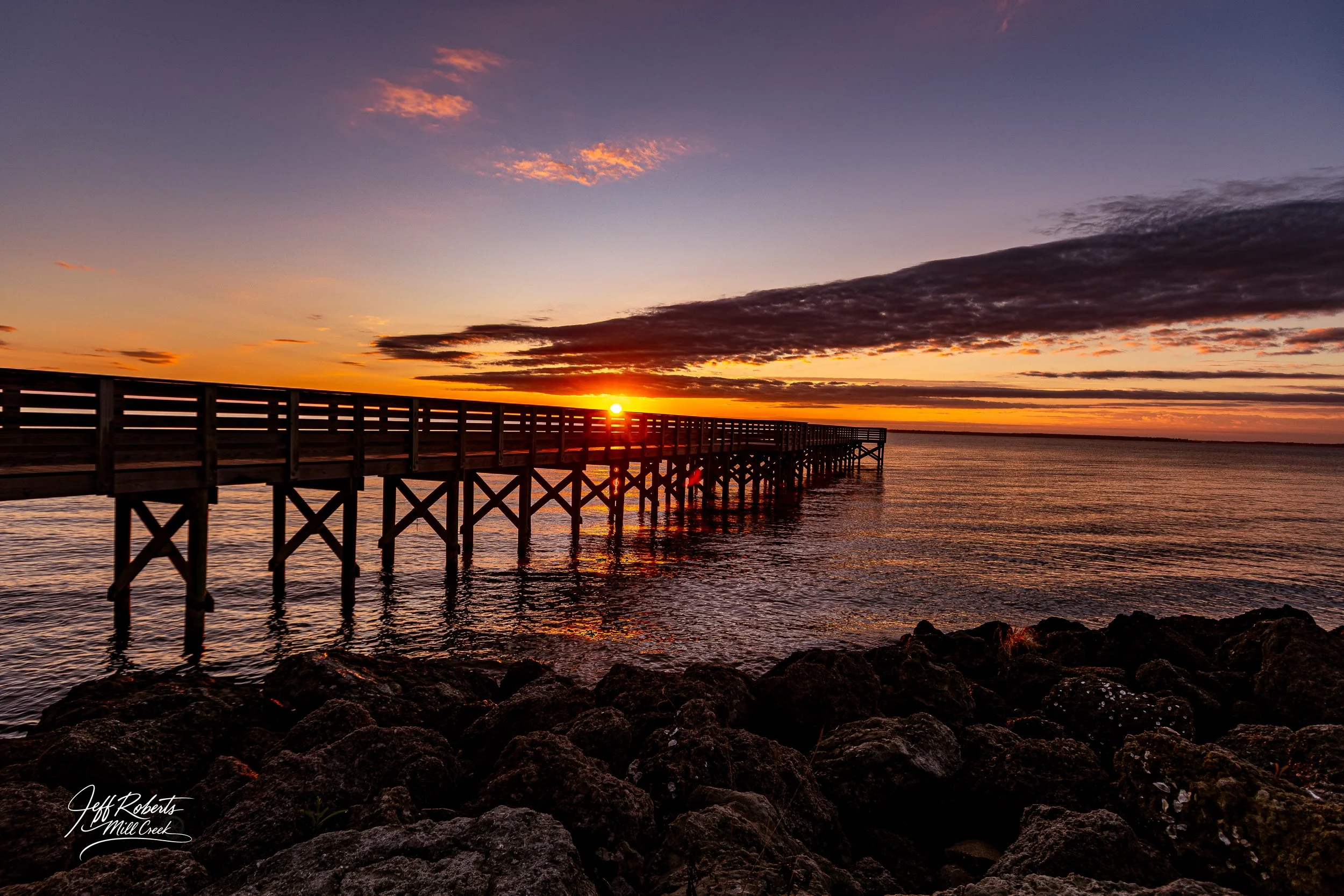 Sunset over a pier extending into a calm body of water, with rocks in the foreground and a partly cloudy sky.