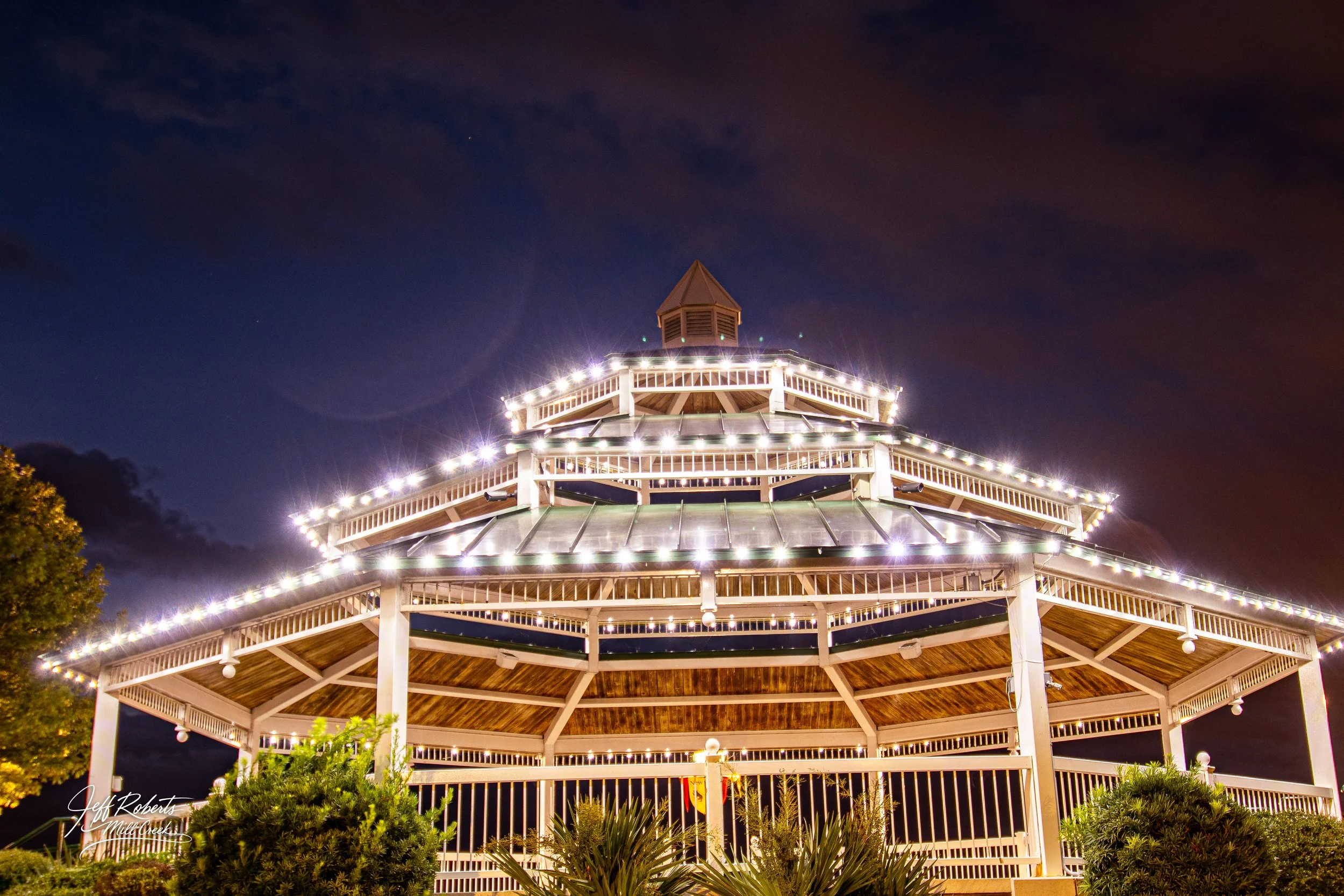 Night view of a multi-tiered, octagonal gazebo decorated with string lights, with a dark sky and some clouds in the background.