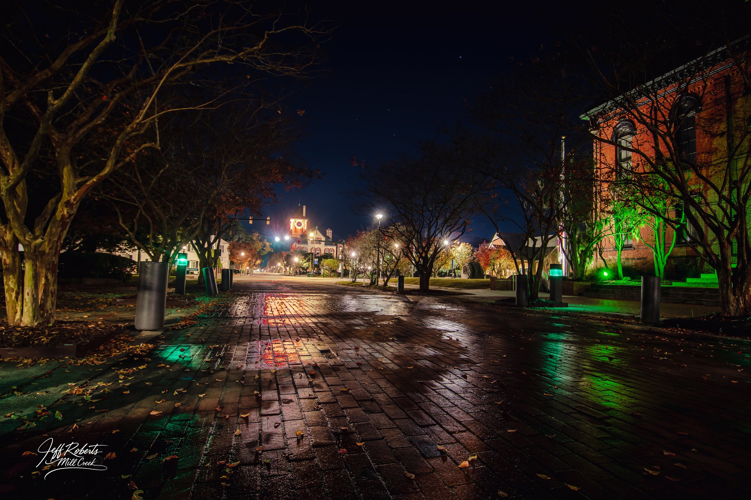Nighttime scene of a wet brick street in downtown Mill Creek, Washington, with trees lining the sidewalk, illuminated buildings, and streetlights reflecting off the wet surface.