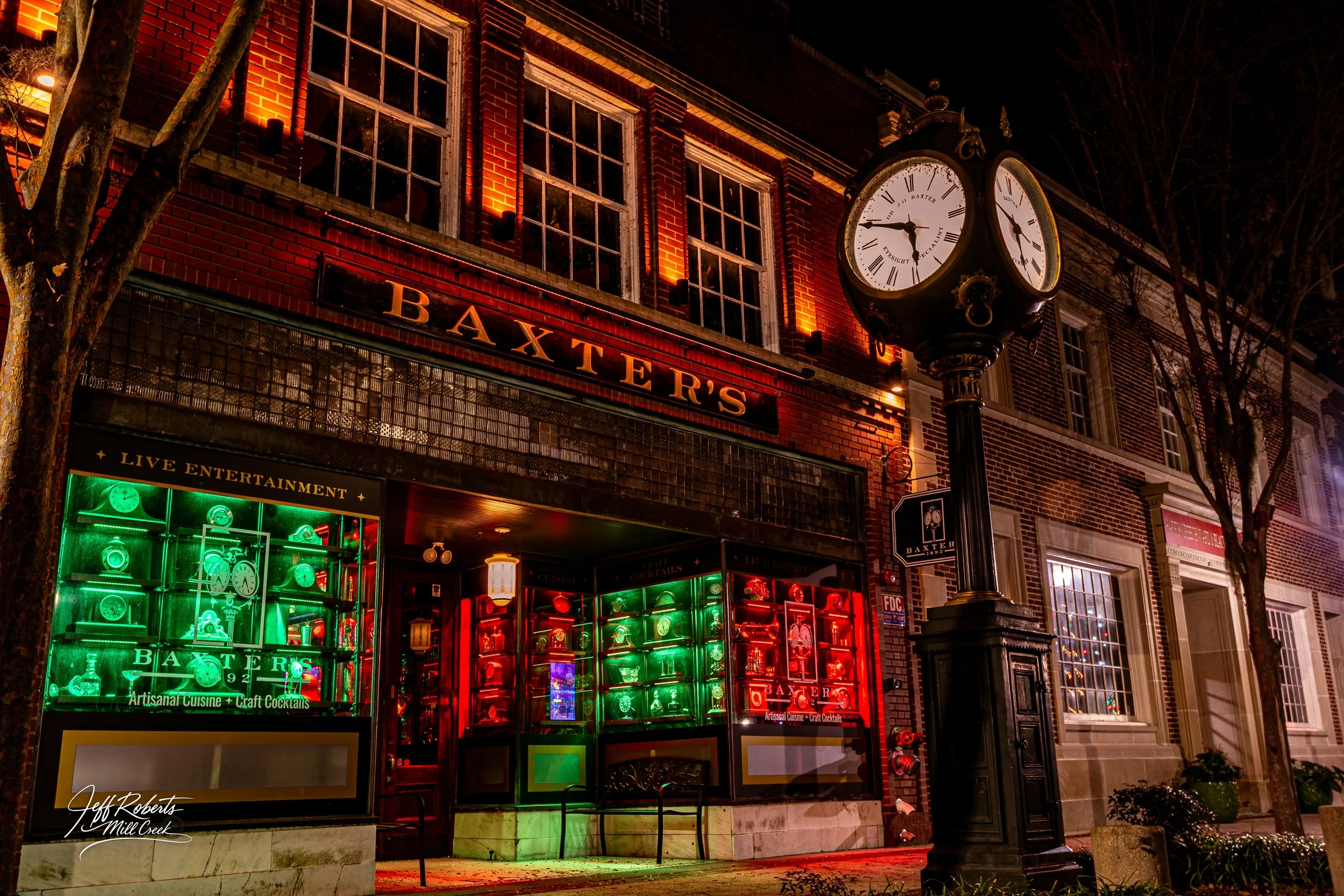 Night view of Baxter's restaurant with a vintage clock on a black street pole, illuminated storefront with green and red neon signs, brick building with lit windows, and bare trees on the sidewalk.