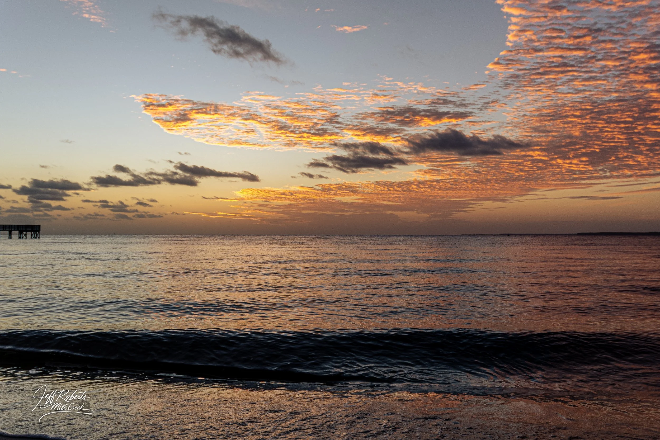 Sunset over the ocean with partly cloudy sky, the clouds illuminated in orange and pink hues, calm water with gentle waves, and a small pier on the left side.