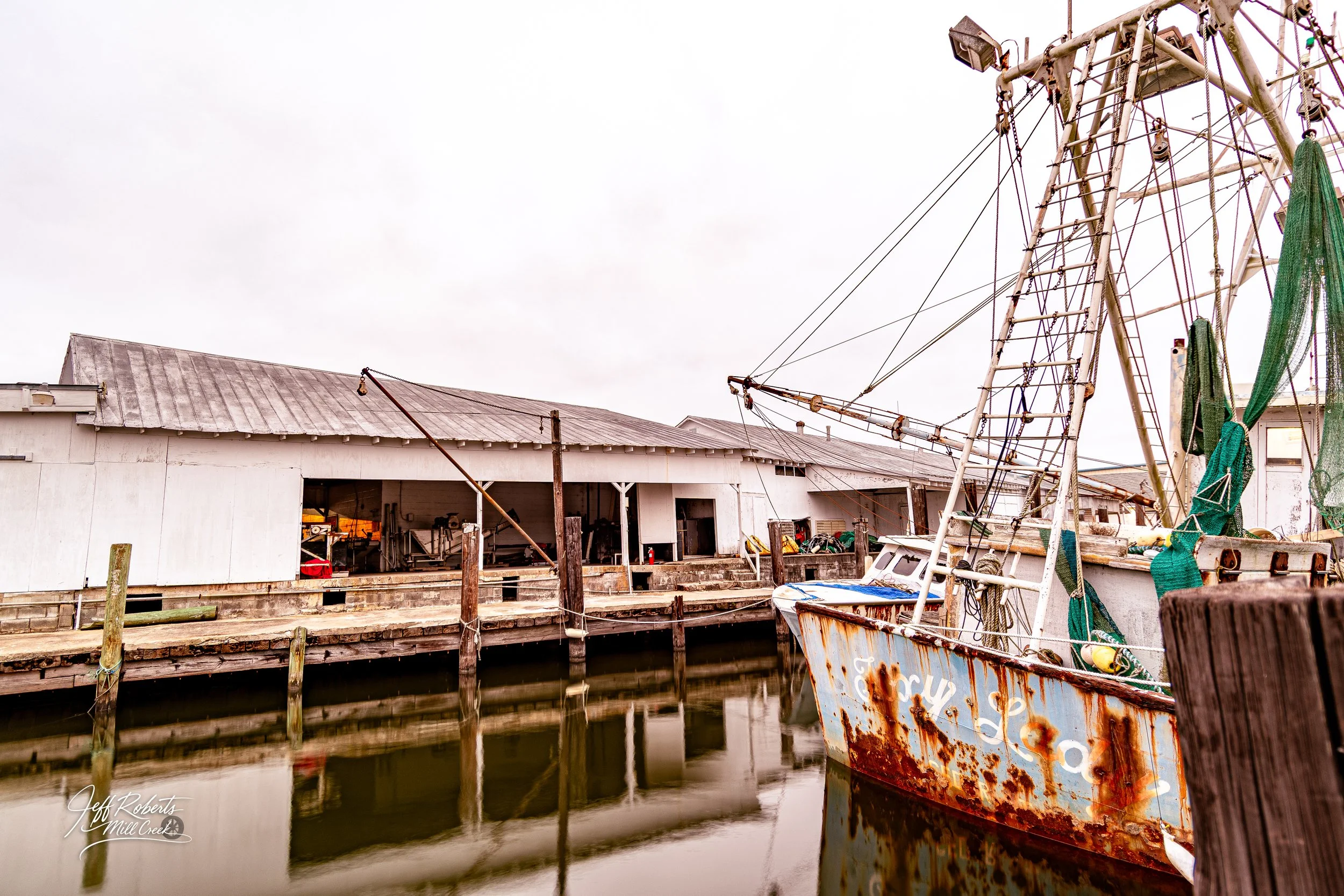 Old rusty fishing boat docked at a wooden pier in front of a weathered boathouse, with water reflecting the scene under an overcast sky.