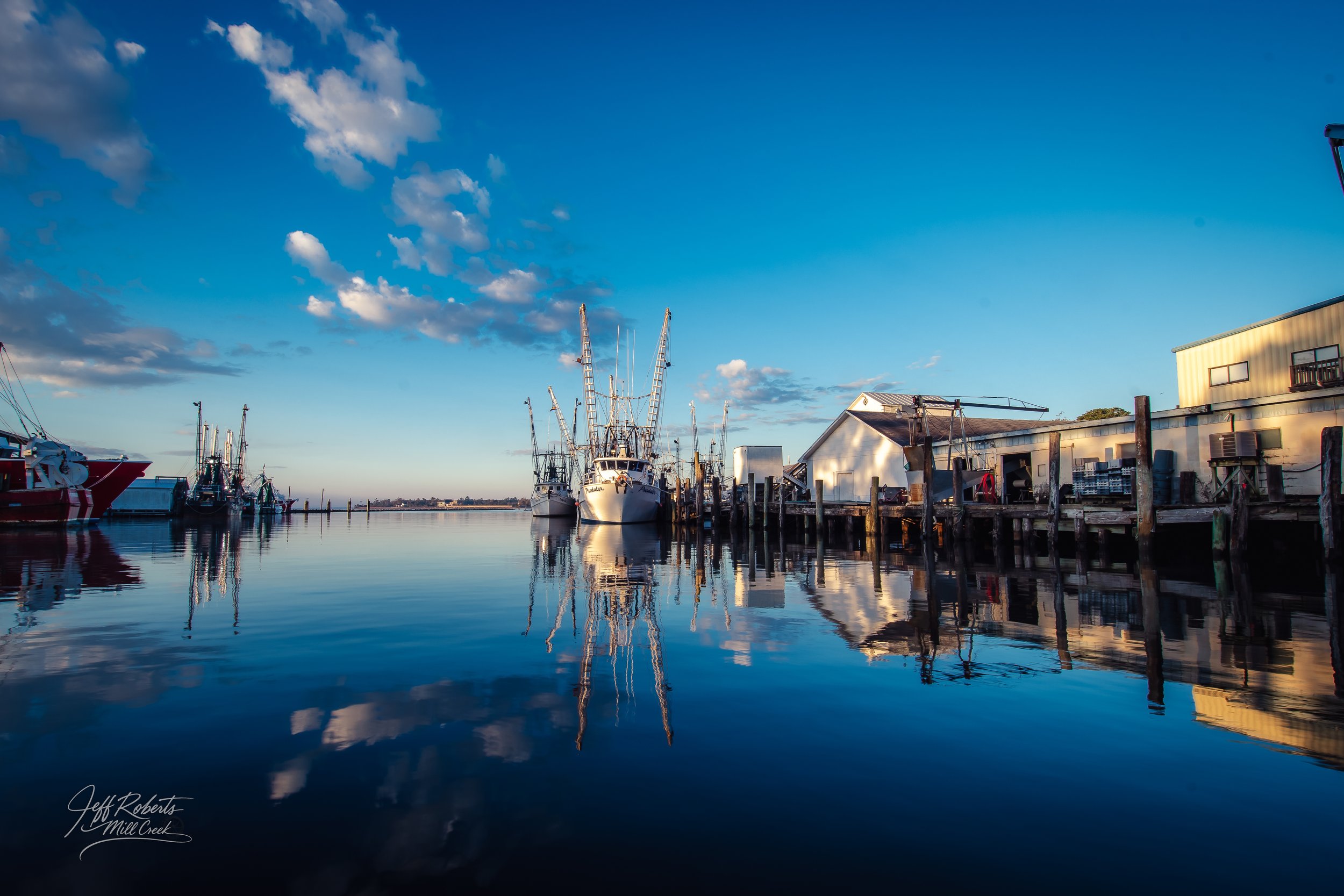 Boats docked at a marina with a clear blue sky and calm water reflecting the vessels and surroundings.