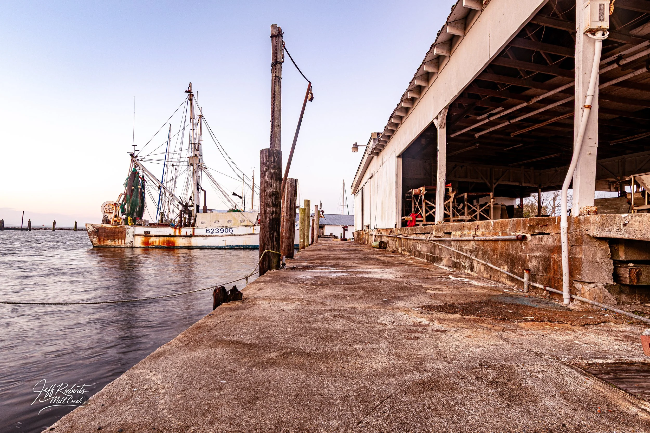 A weathered boat dock with a large white boat moored on the left and a wooden shed or boathouse on the right. The dock is made of concrete with posts and ropes securing the boat, and the sky is clear with soft lighting.