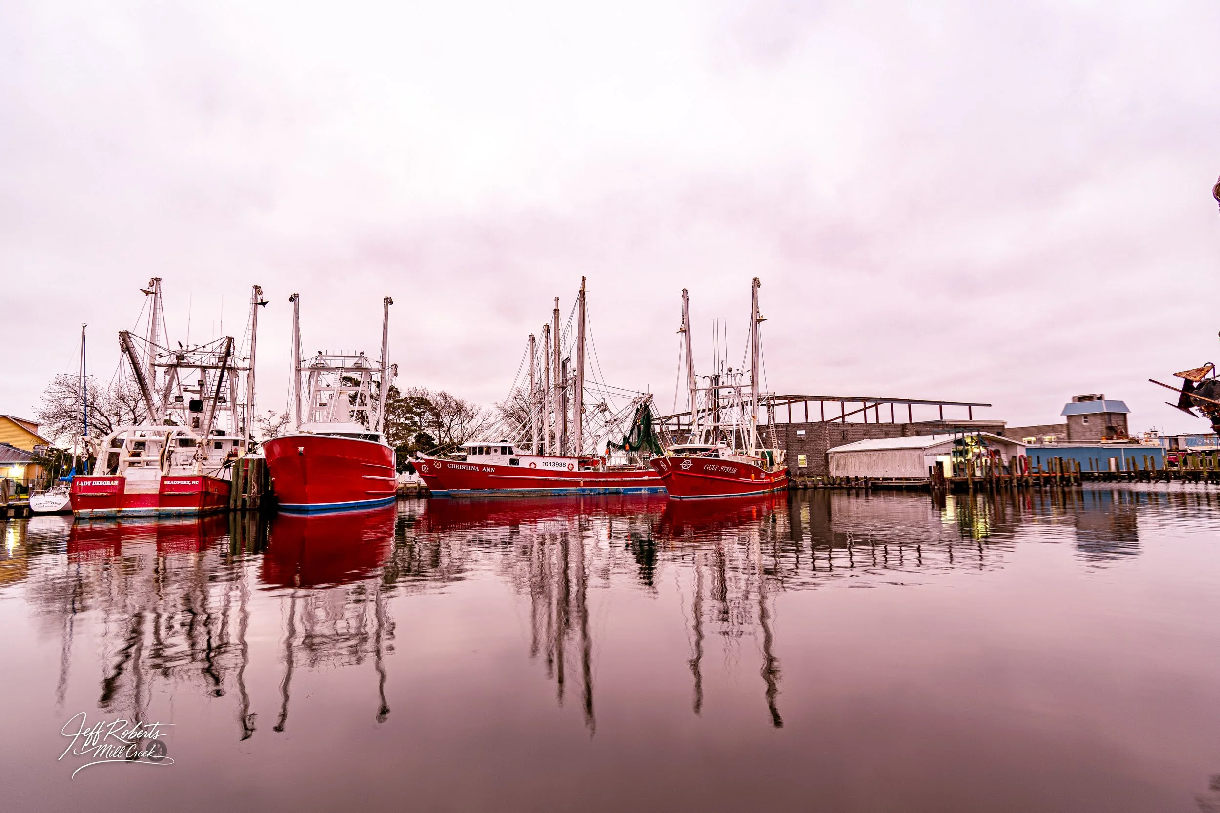 Several red and white boats docked at a marina with reflections on the water, overcast sky, and buildings along the shoreline in the background.