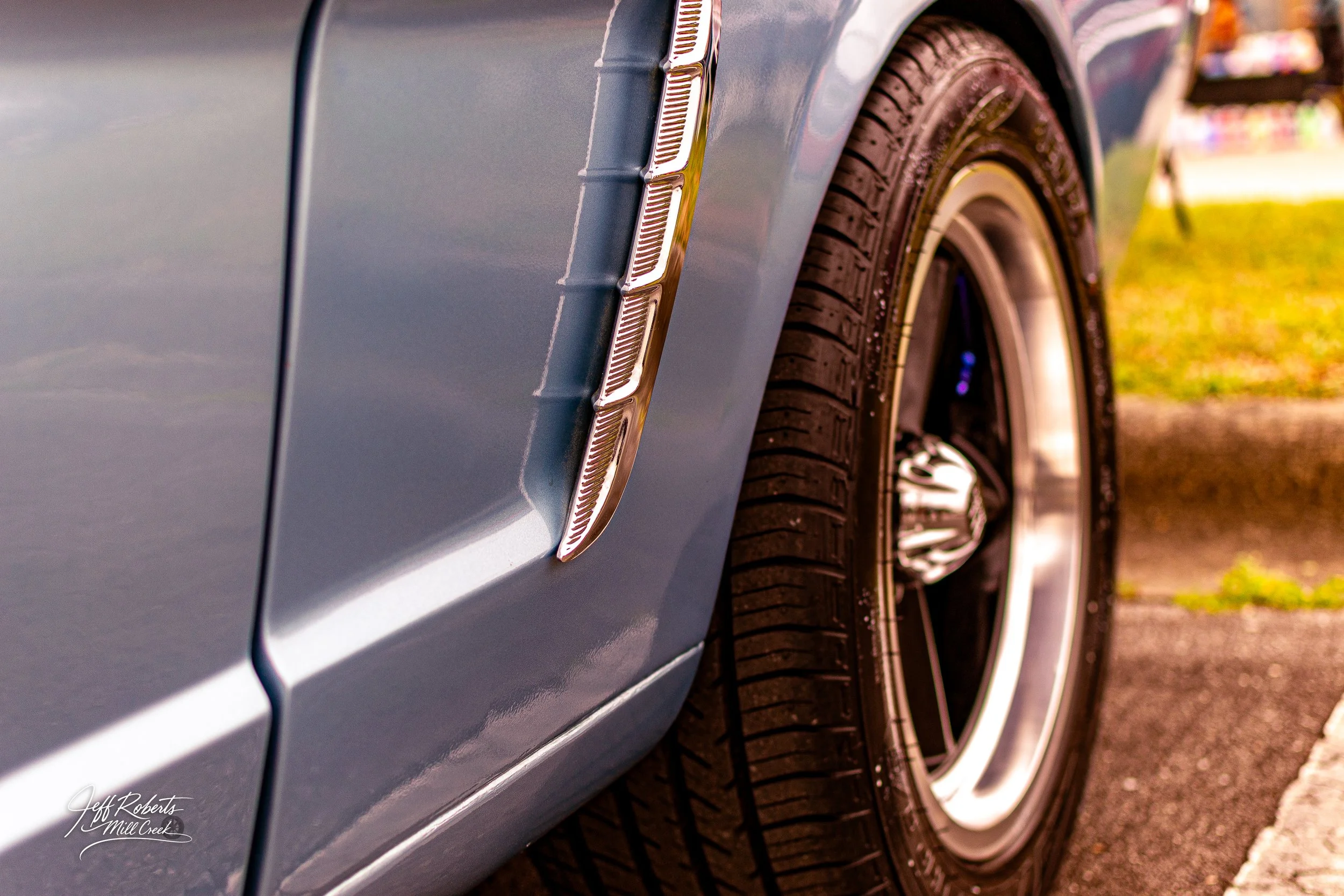 Close-up of a classic car's front fender, showing a chrome accent and black tire with a shiny hubcap, on pavement with grass in the background.