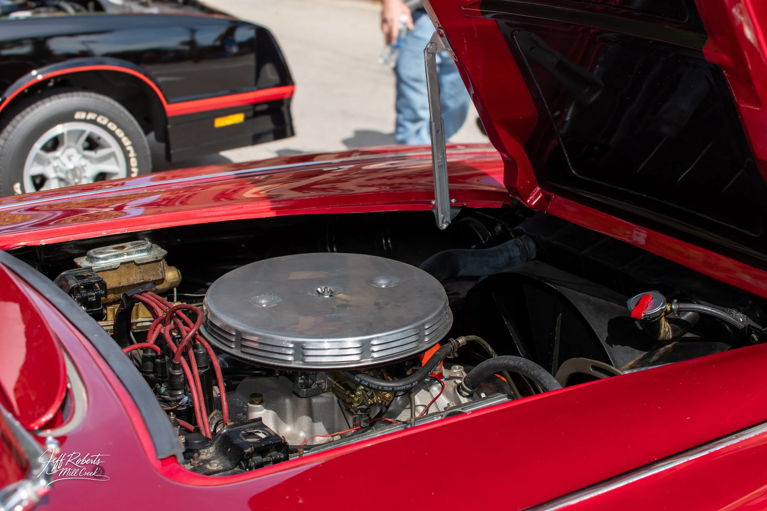 Close-up of a red vintage car's open hood revealing the engine with a silver air cleaner and red spark plug wires, at a car event with a black and red classic car in the background.