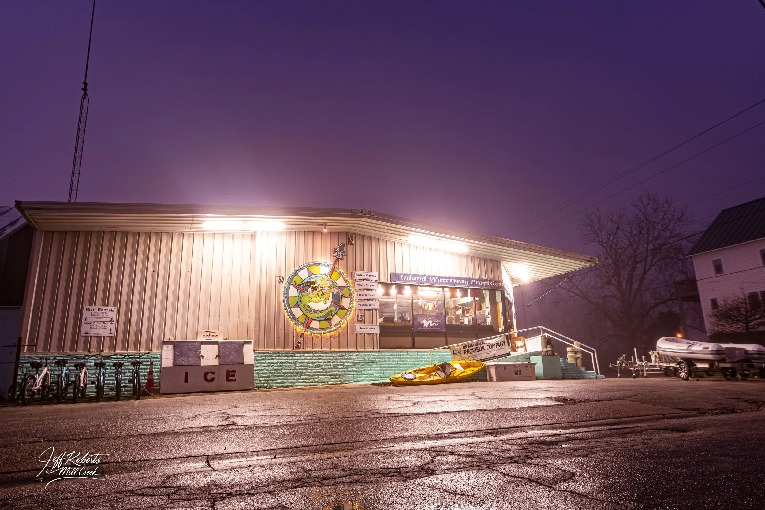 Nighttime view of a waterway rental shop with colorful signage and a nautical dragon logo, illuminated by exterior lights, with a kayak on the ground, a few boats on trailers, and a sign indicating ice for sale, in a small town with wire power lines 
