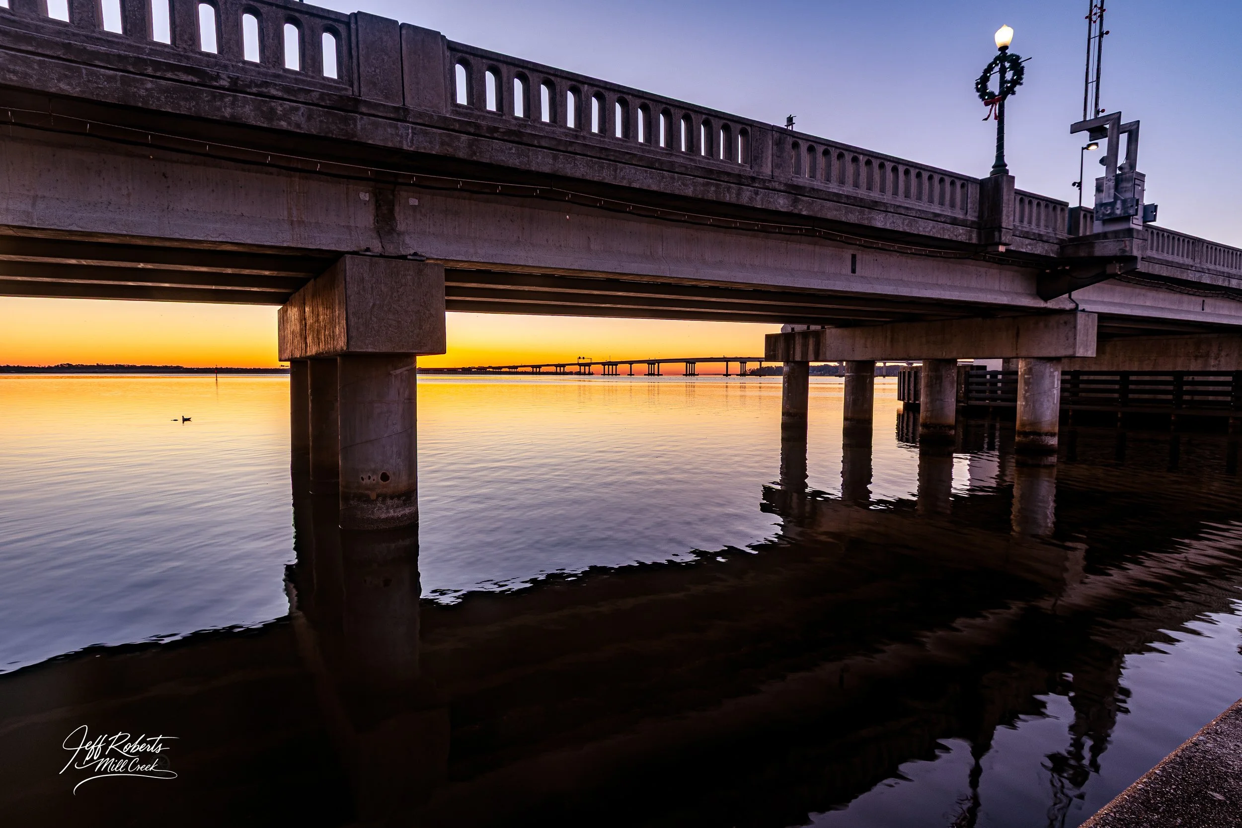 A concrete bridge over a calm river at sunset with soft orange and purple sky, reflection in water, and streetlamp with holiday wreath on top.