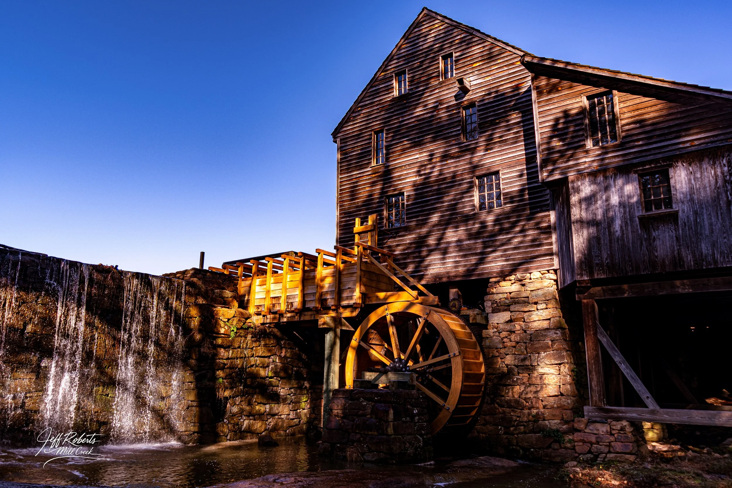 Historic wooden mill with waterwheel beside a waterfall under a clear blue sky.
