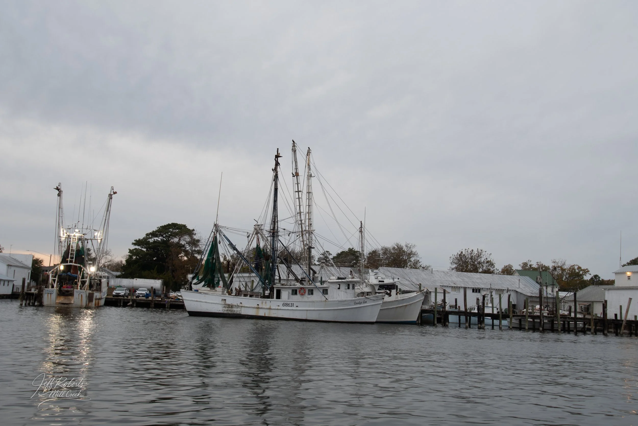 Two boats docked at a marina with overcast sky and wooden pier, white buildings in background.