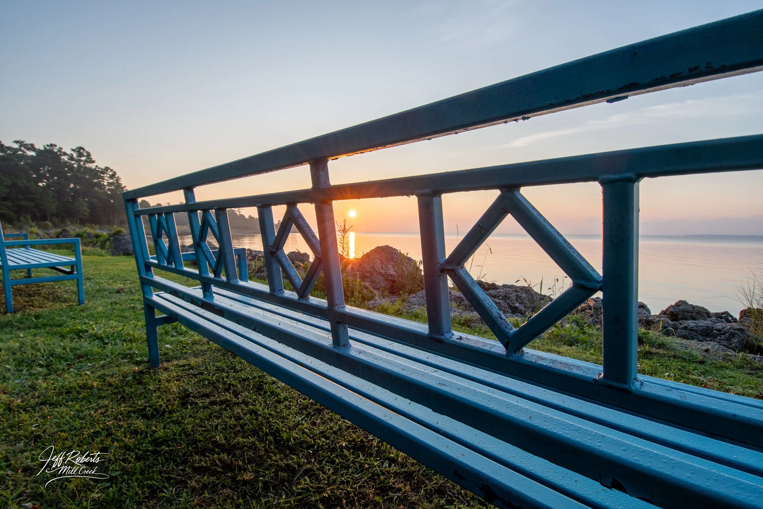 A blue park bench and railing overlooking a body of water during sunset, with rocks and trees in the background.