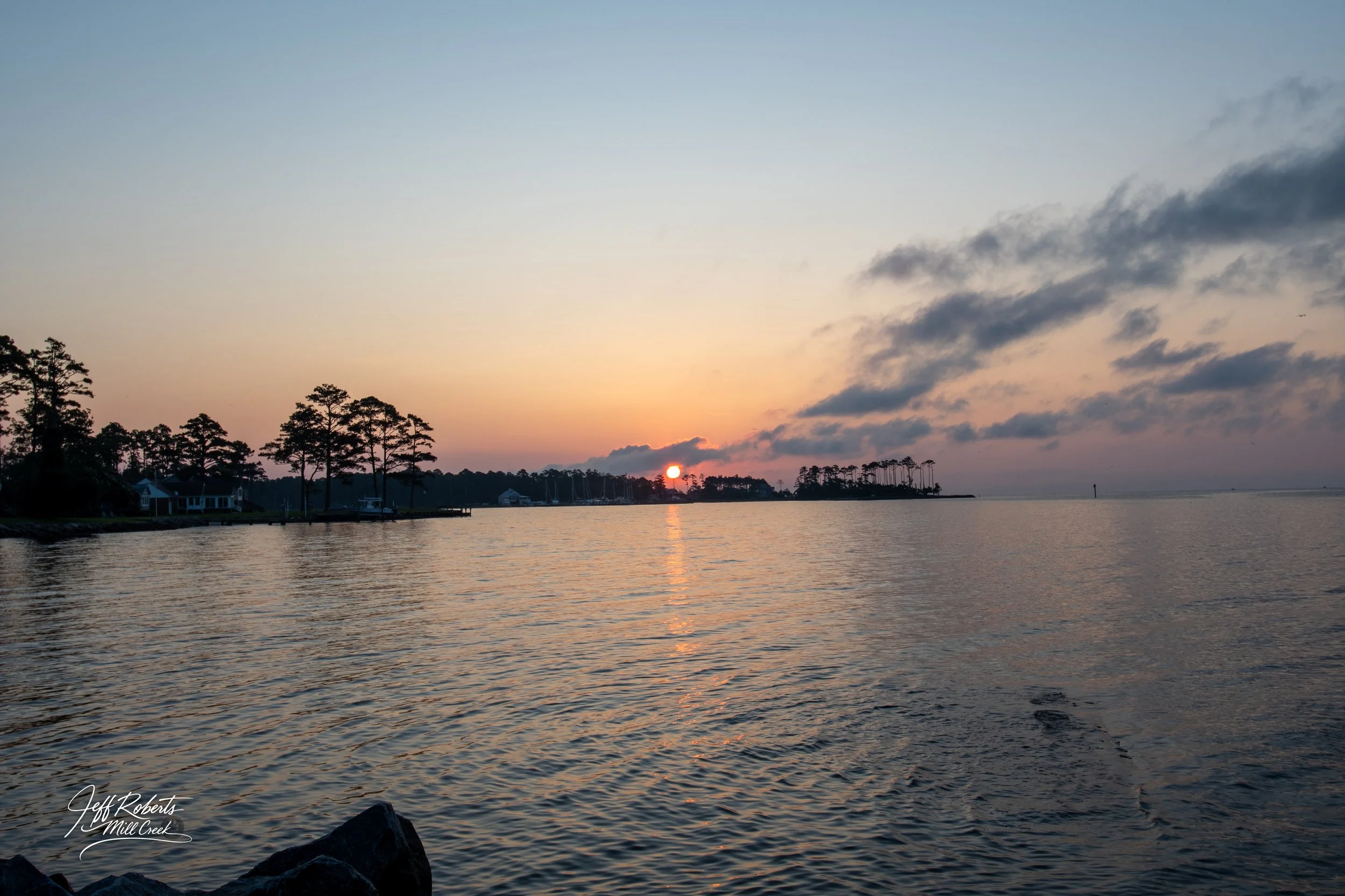 Sunset over a calm body of water with trees on the shoreline and a partly cloudy sky.