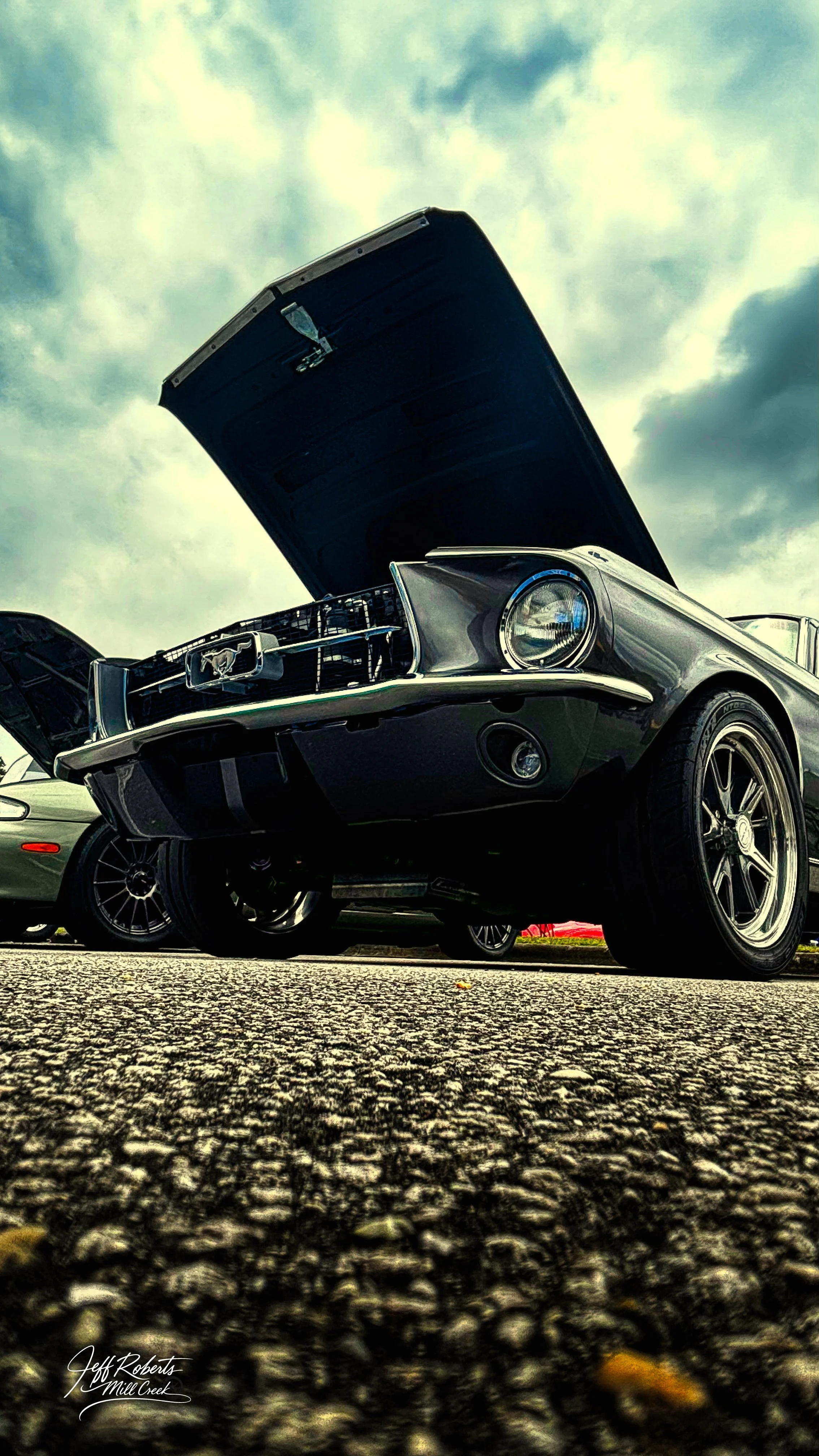 A vintage black Ford Mustang with its hood open, parked on a gravel surface under a cloudy sky.