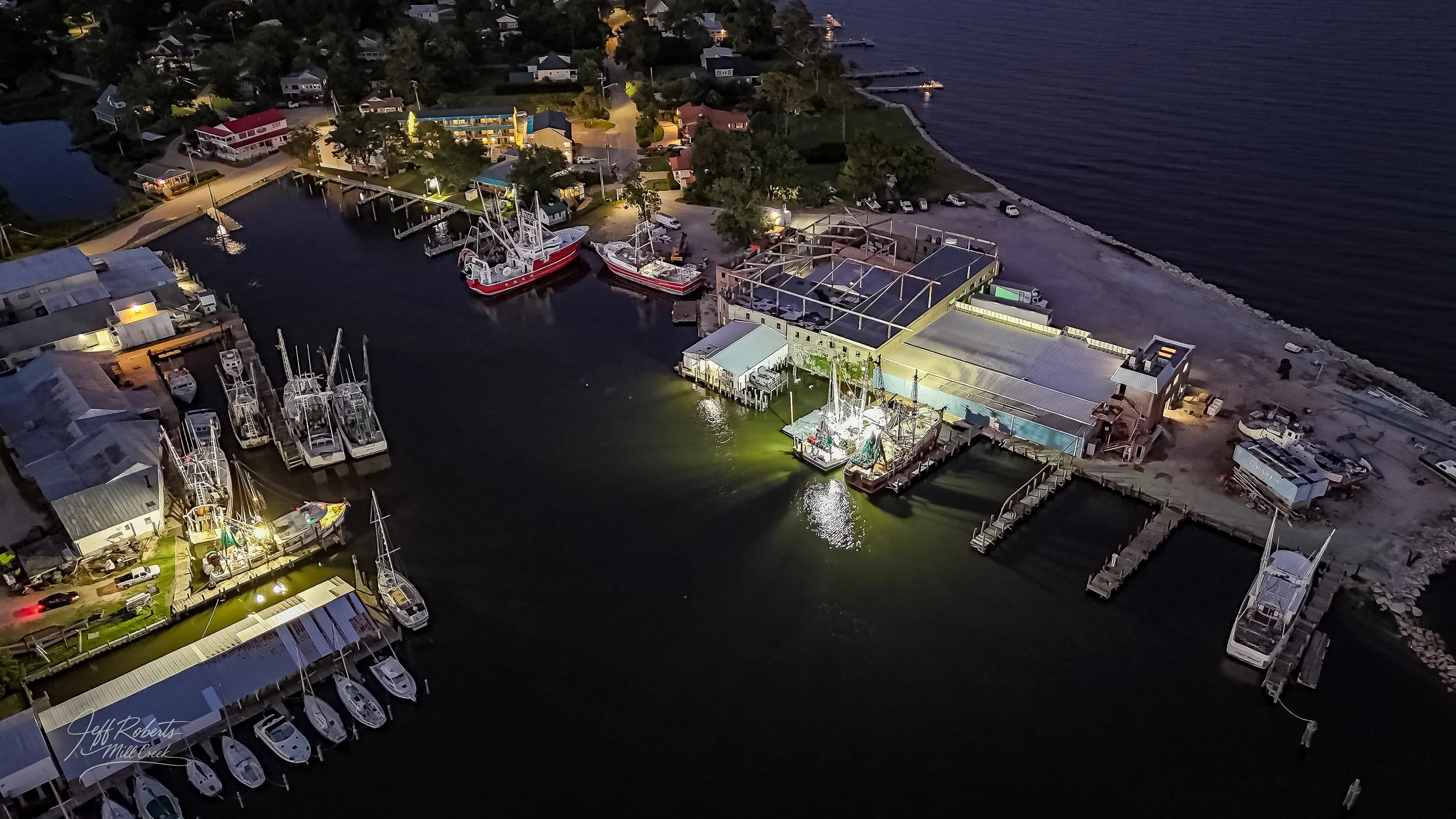 An aerial view of a marina at night with boats docked at piers, surrounded by buildings and a residential area, illuminated by outdoor lights.