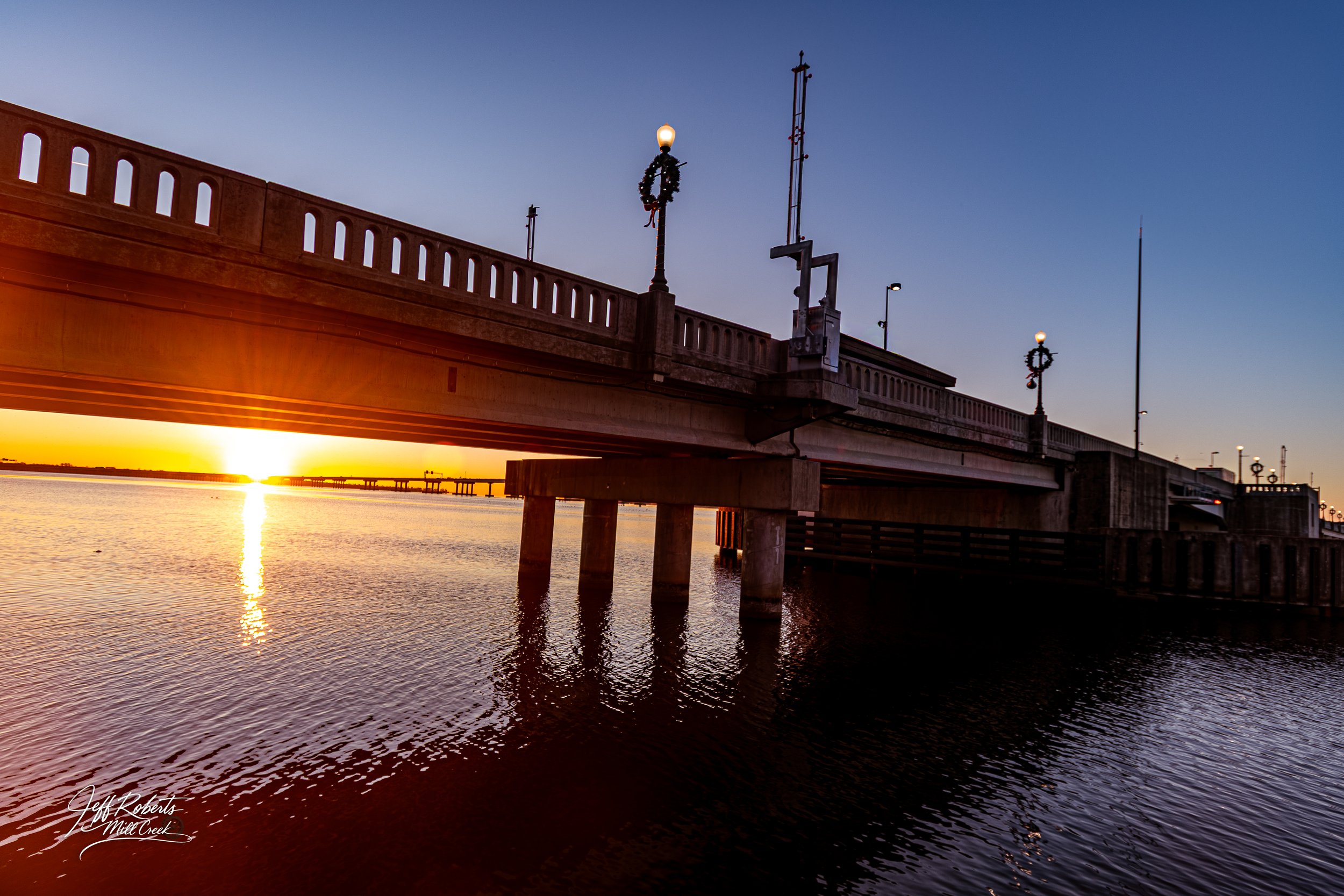 Sunset over a river with a bridge in the foreground, decorated with Christmas wreaths and streetlights.