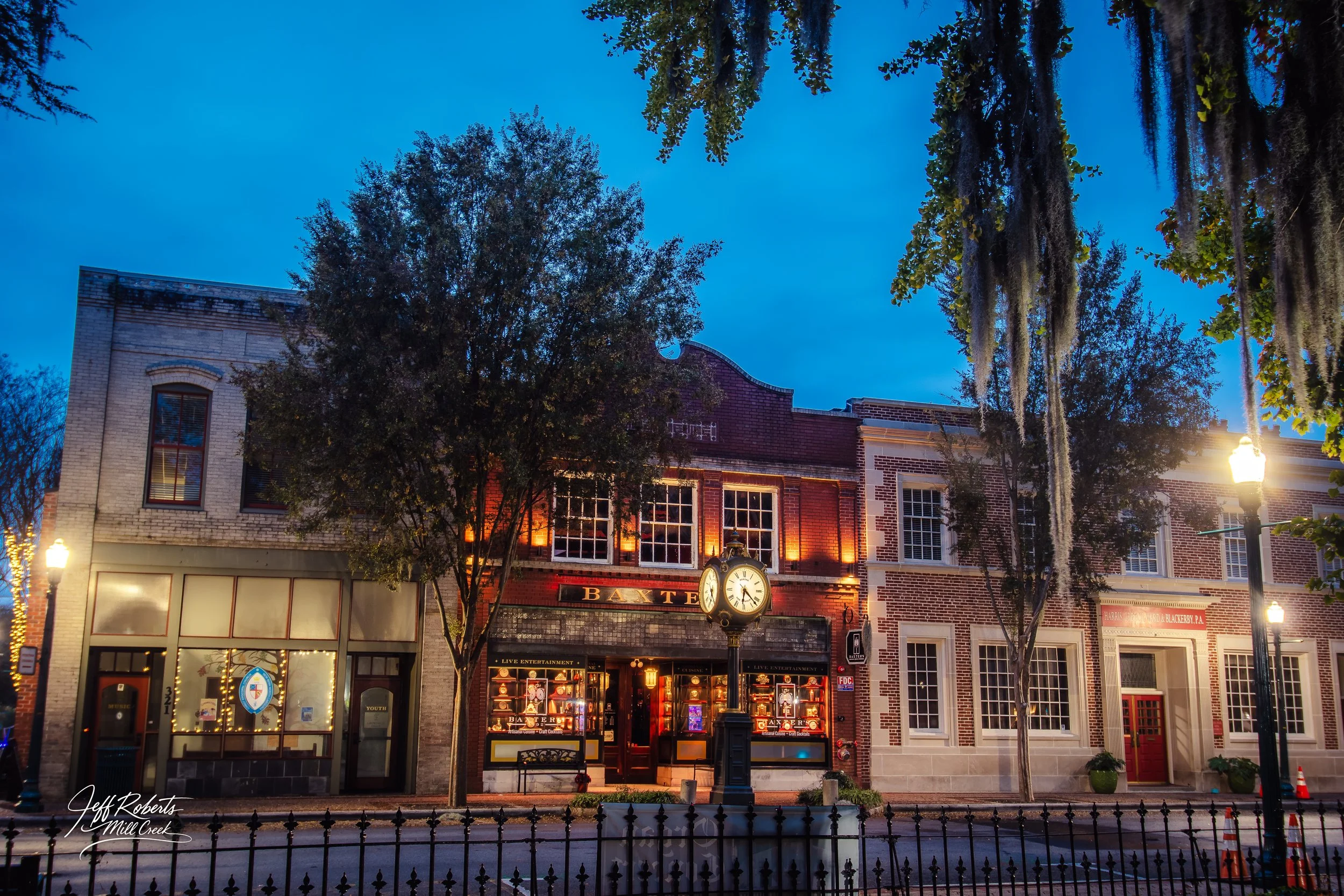 Historic brick building with a large clock in front, illuminated by streetlights at dusk, with trees and a black iron fence in the foreground.