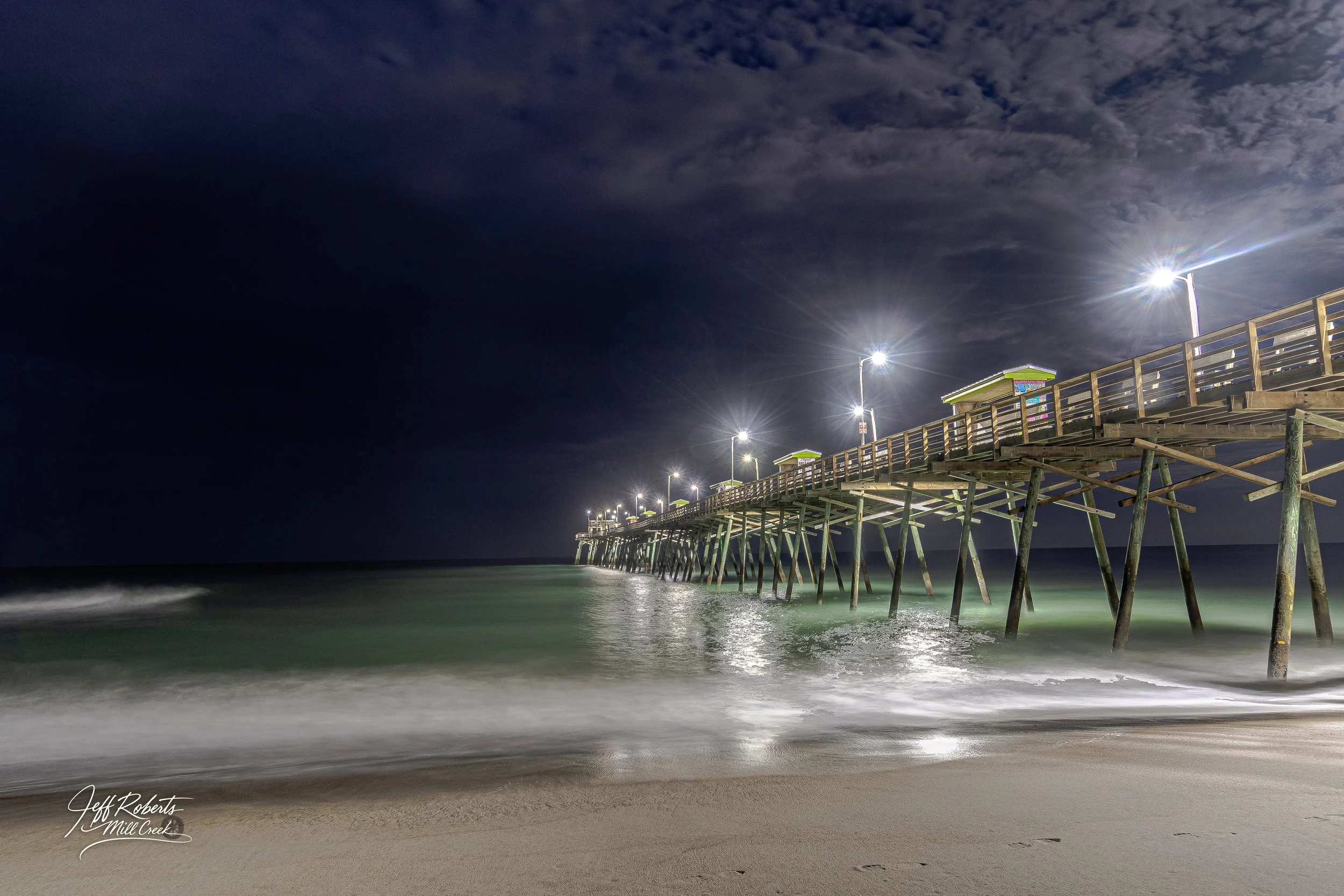 Nighttime view of a wooden pier extending into the ocean with bright lights along its length, under a dark sky with some clouds.