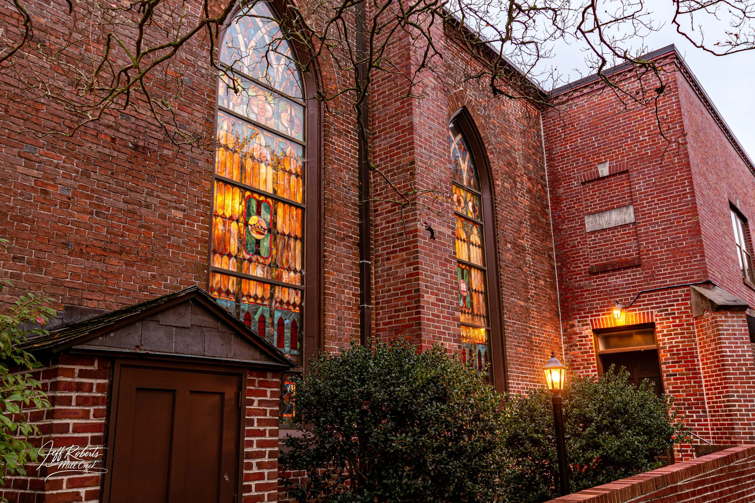 A brick church with tall stained glass windows showing vibrant colors and religious motifs. Small bushes in front and a lit streetlamp near the entrance.