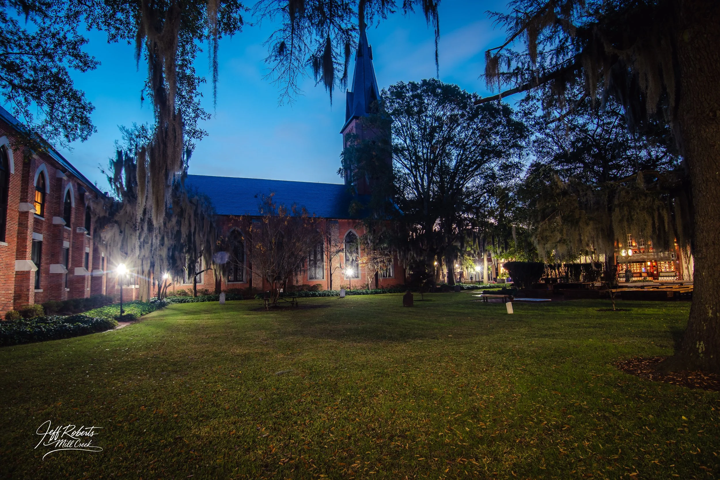 Night view of a red brick church with a tall steeple, surrounded by trees with Spanish moss, lit by outdoor lights, with a grass lawn in the foreground.