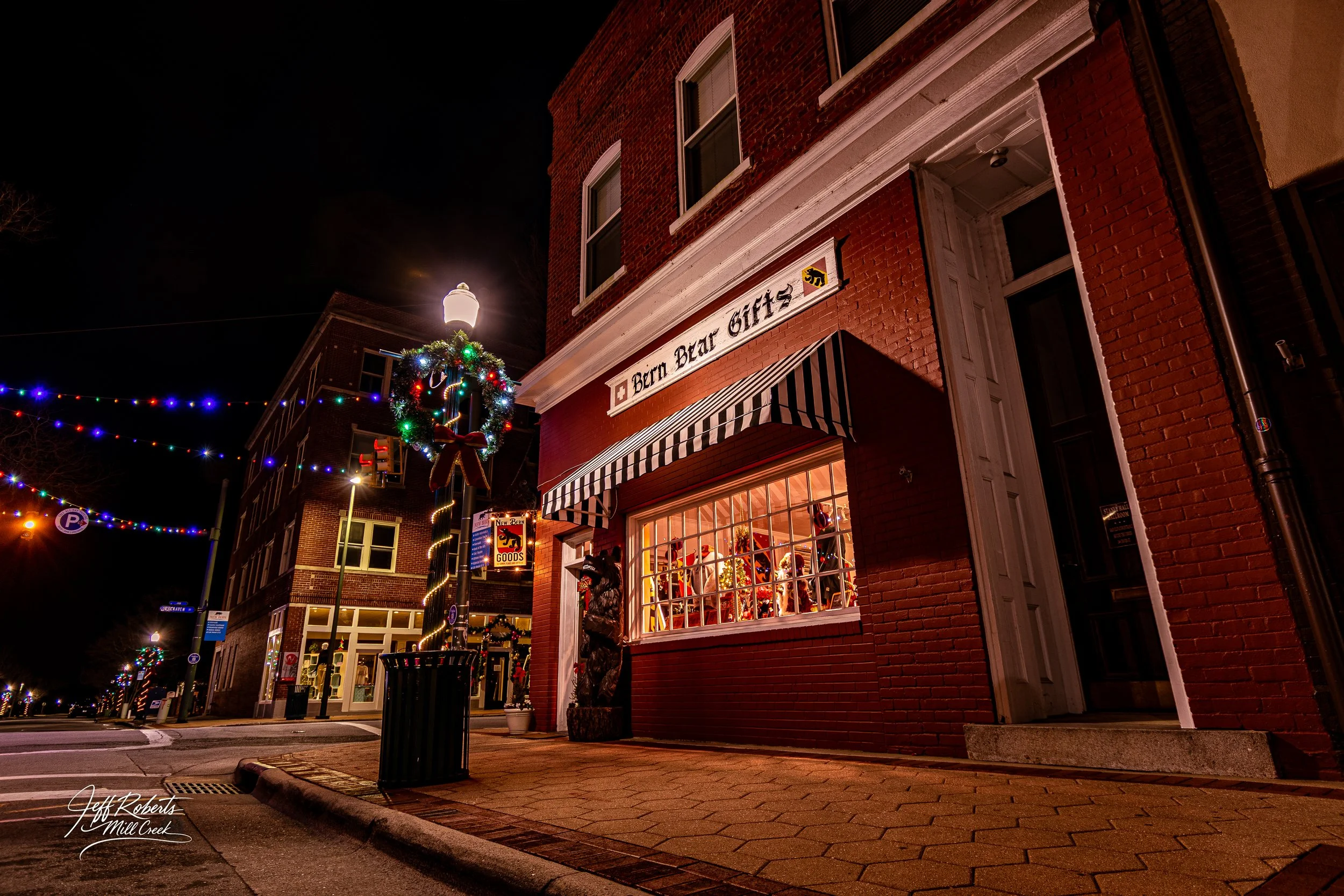 Nighttime street scene featuring a decorated Christmas wreath on a lamppost, with a vintage brick building displaying a gift shop called 'Berr Bear Gifts.' The shop window is lit up with holiday decorations, and multicolored string lights span across