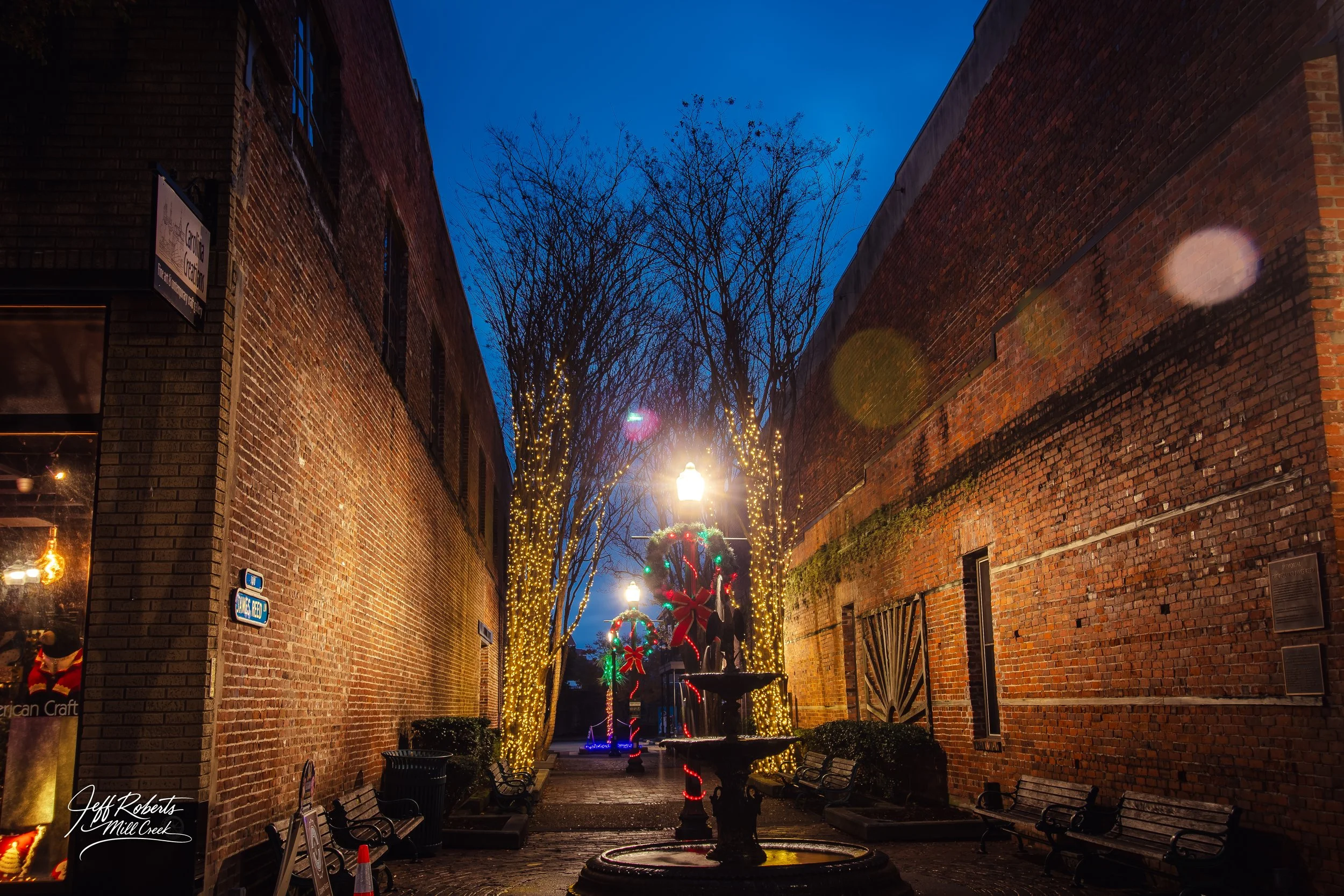 A narrow alleyway with brick walls on both sides decorated with Christmas lights, featuring a fountain and benches, with Christmas wreaths and lighted trees in the background, under a dark blue evening sky.