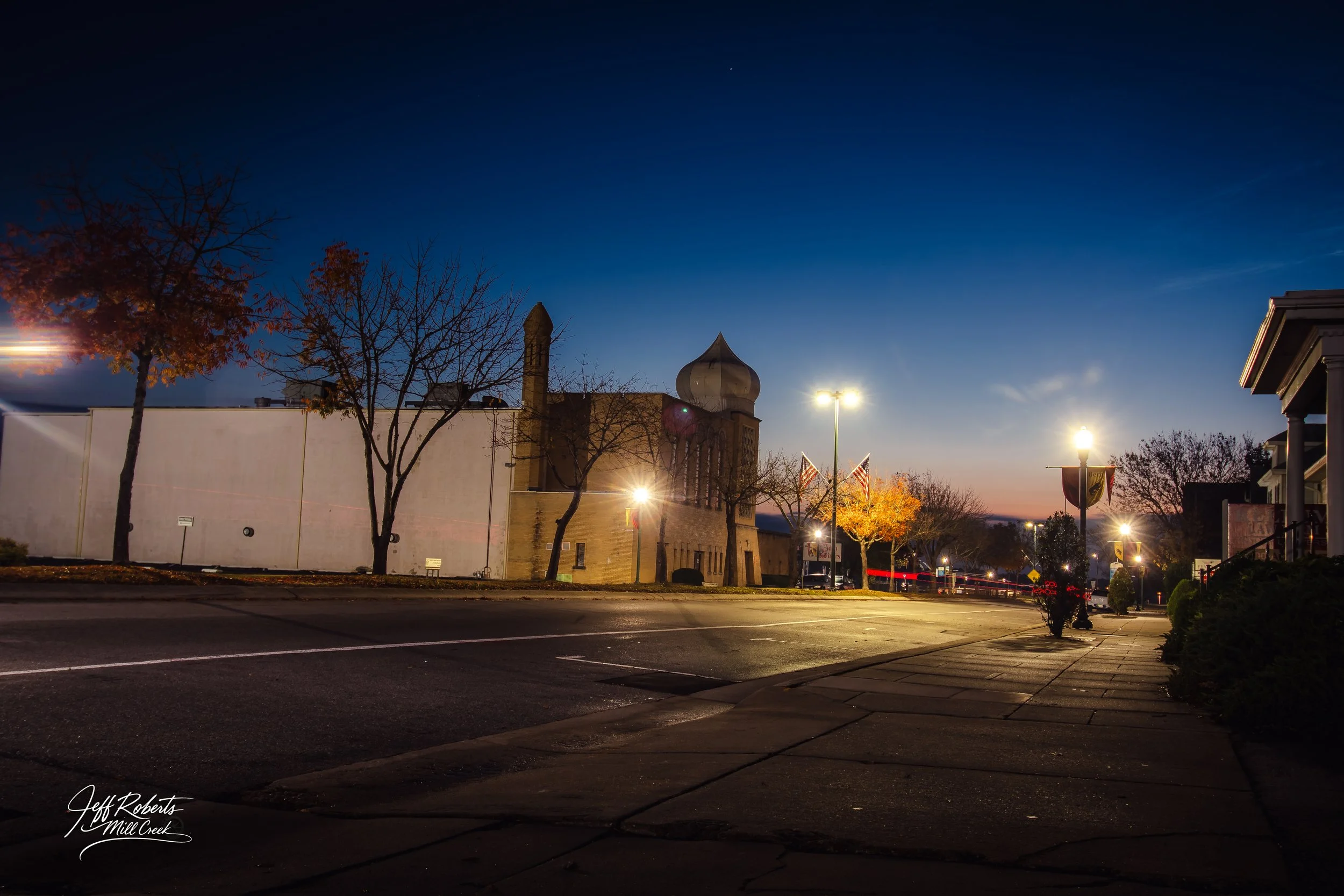 A quiet city street at dusk with streetlights illuminating the sidewalk, leafless trees, and a historic building with a dome and tower in the background.