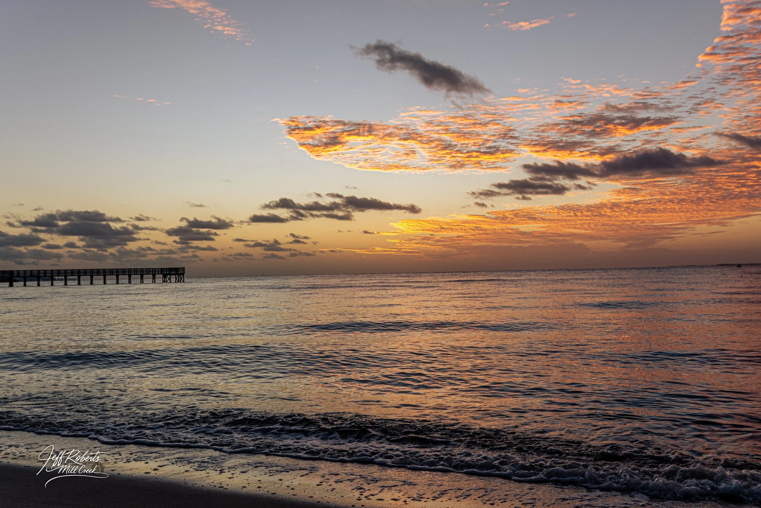 Sunset over the ocean with partly cloudy sky, small waves on the shore, and a pier extending into the water on the left side.