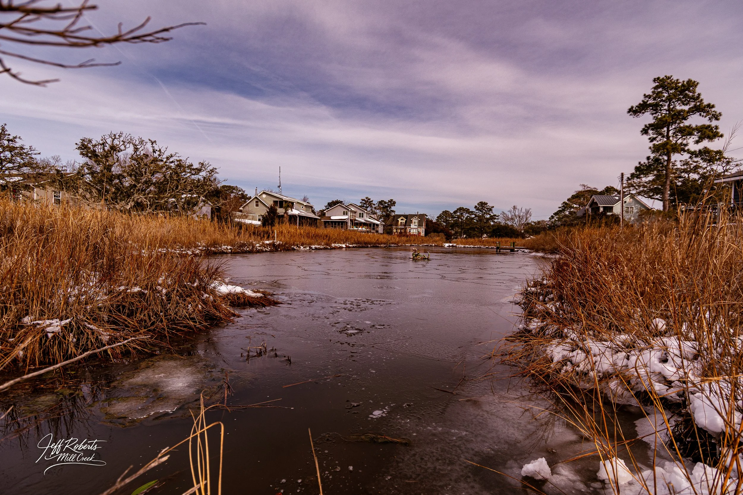 A partially frozen creek with tall, brown grasses lining its banks, snow remnants on the ground, and houses in the background under a partly cloudy sky.