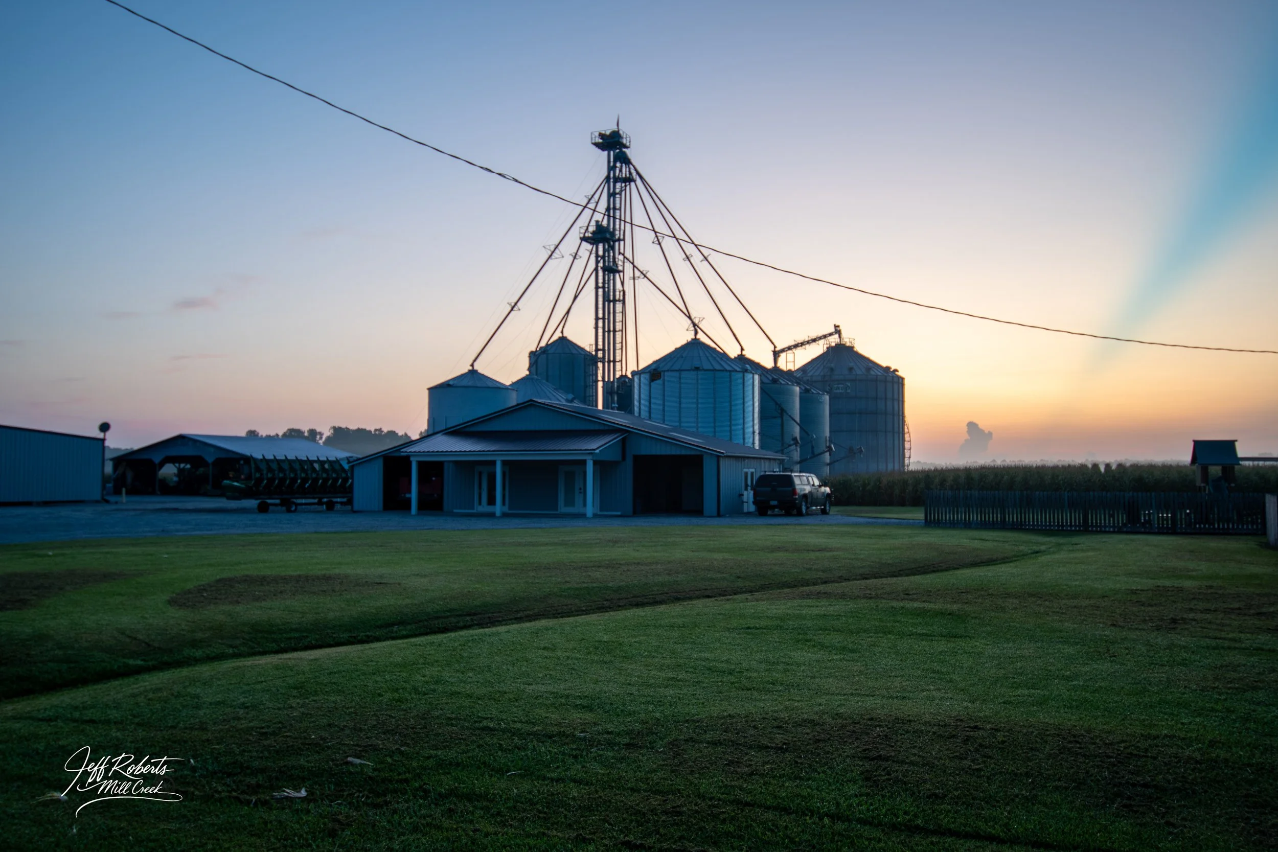 A farm at sunset with silos, a barn, and a field of grass.