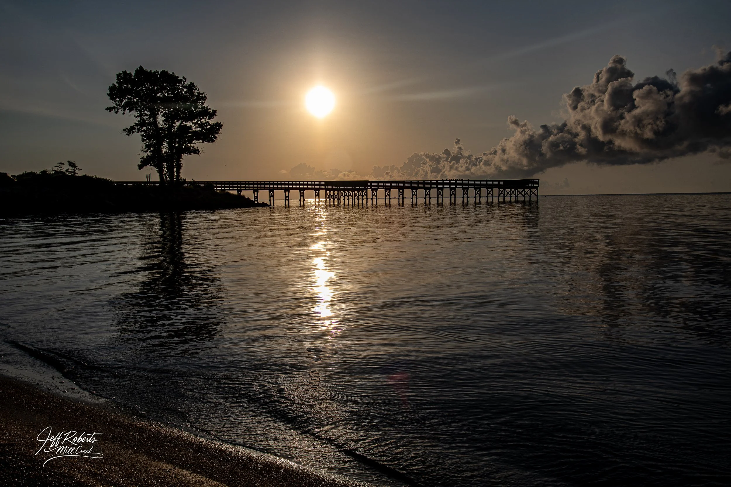 Sunset over a waterbody with a wooden pier extending into the distance, a tree on the left, reflections on the water, and clouds in the sky.