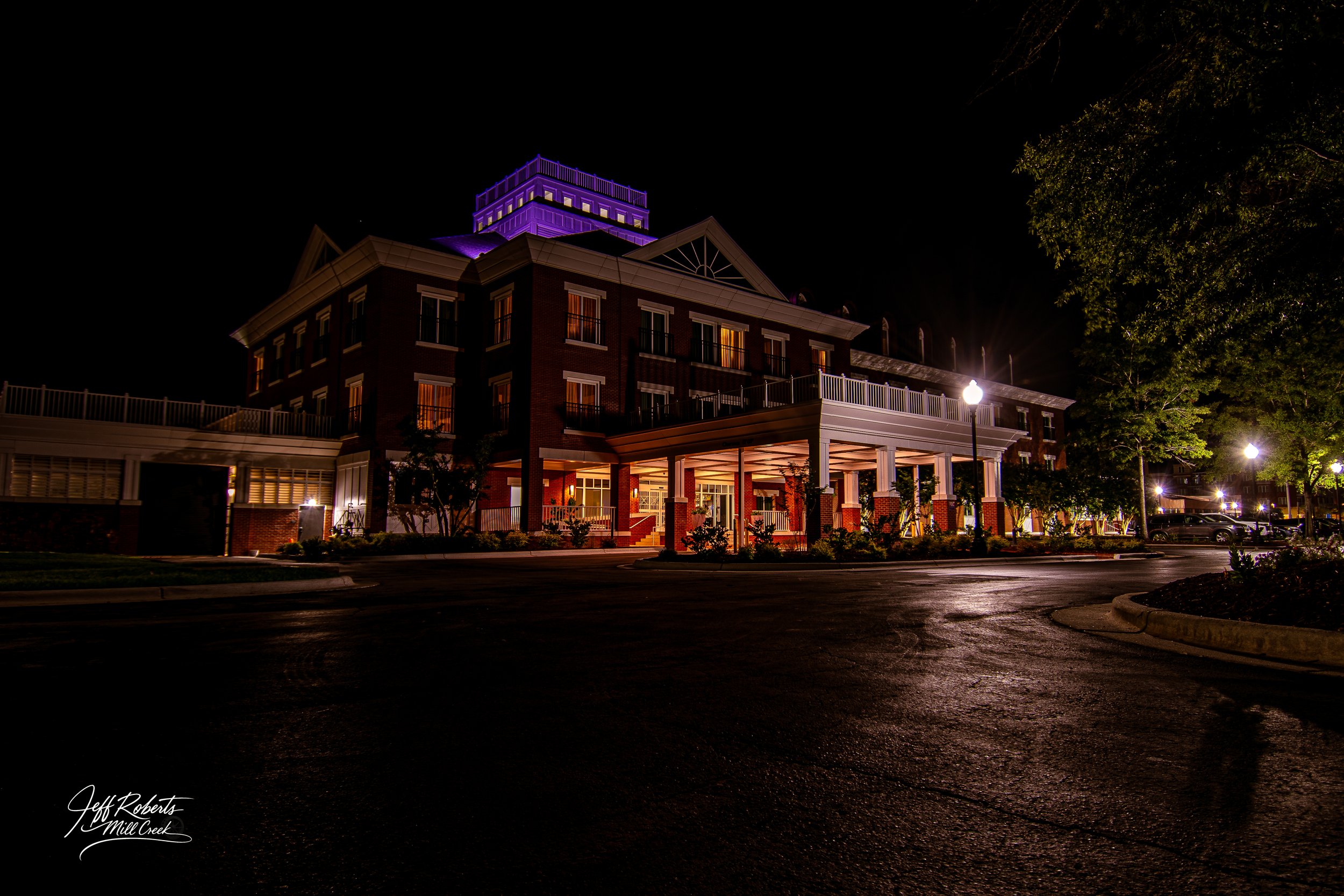 Nighttime exterior view of a multi-story building with warm interior lighting, surrounded by street lamps and trees, with a dark sky in the background.